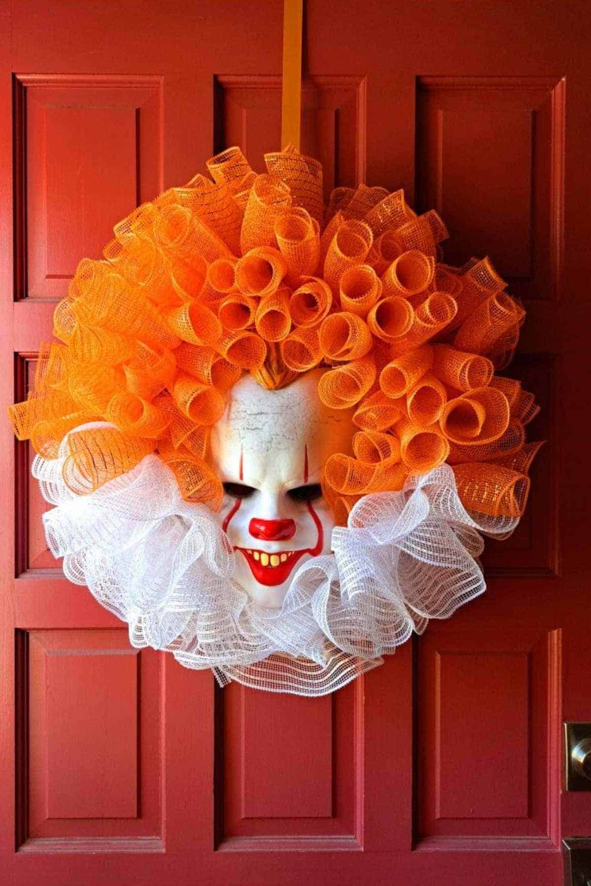 A clown mask wreath with orange and white mesh decorations hangs on a red door.