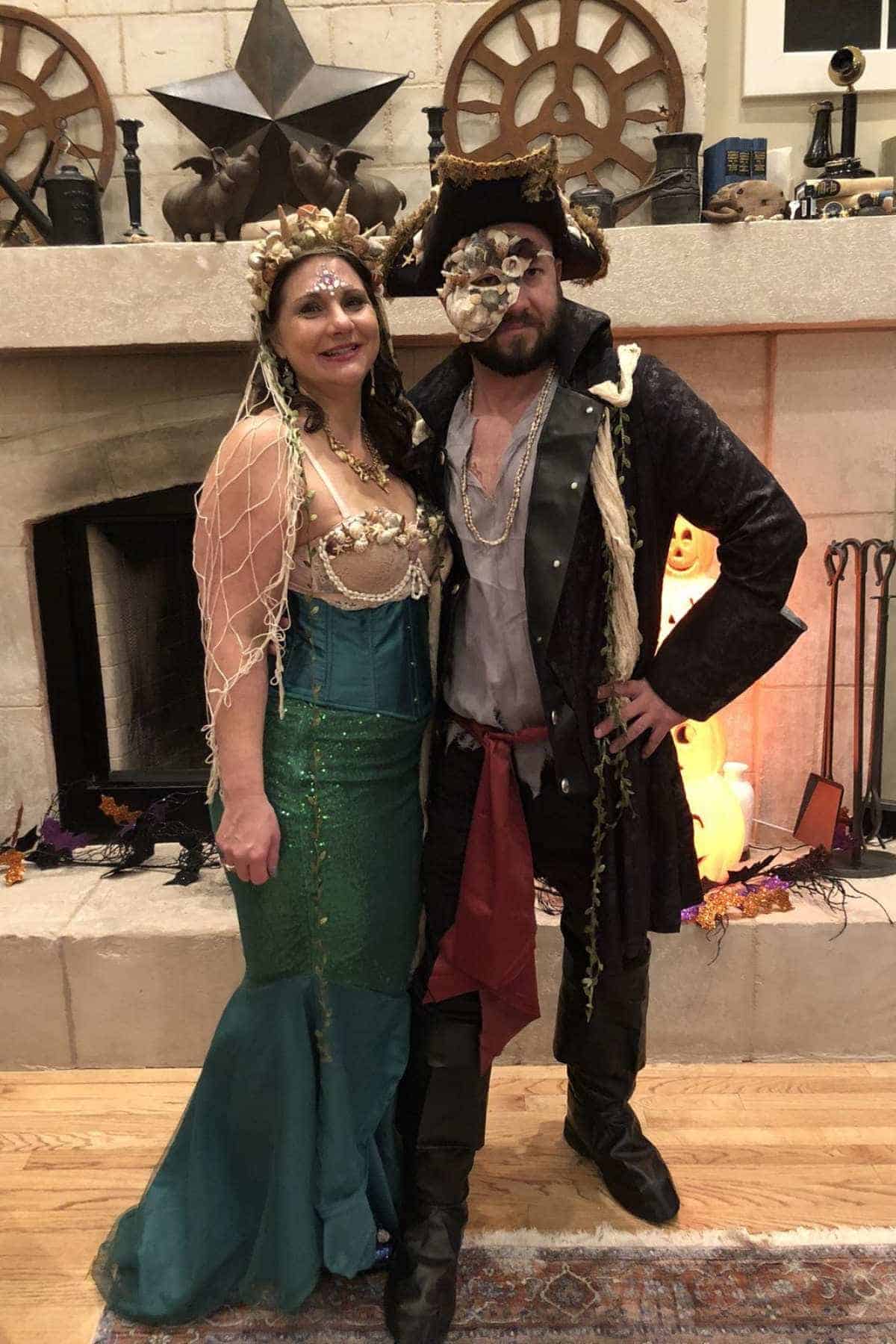 A woman in a mermaid costume stands next to a man in a pirate outfit with an eye patch and hat, posing indoors in front of a decorated fireplace.