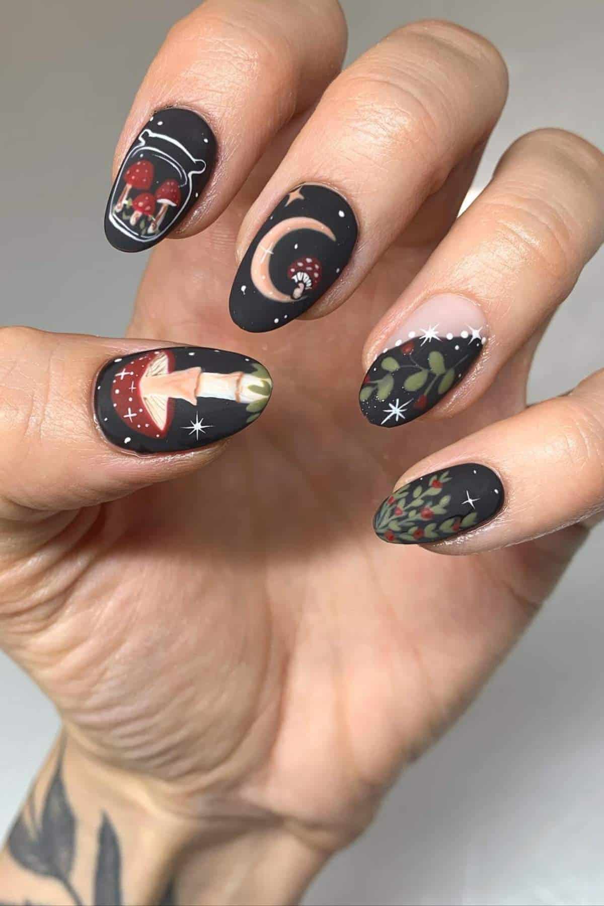 A hand with black matte nail art featuring mushrooms, stars, a crescent moon, and leafy designs, held against a plain background.