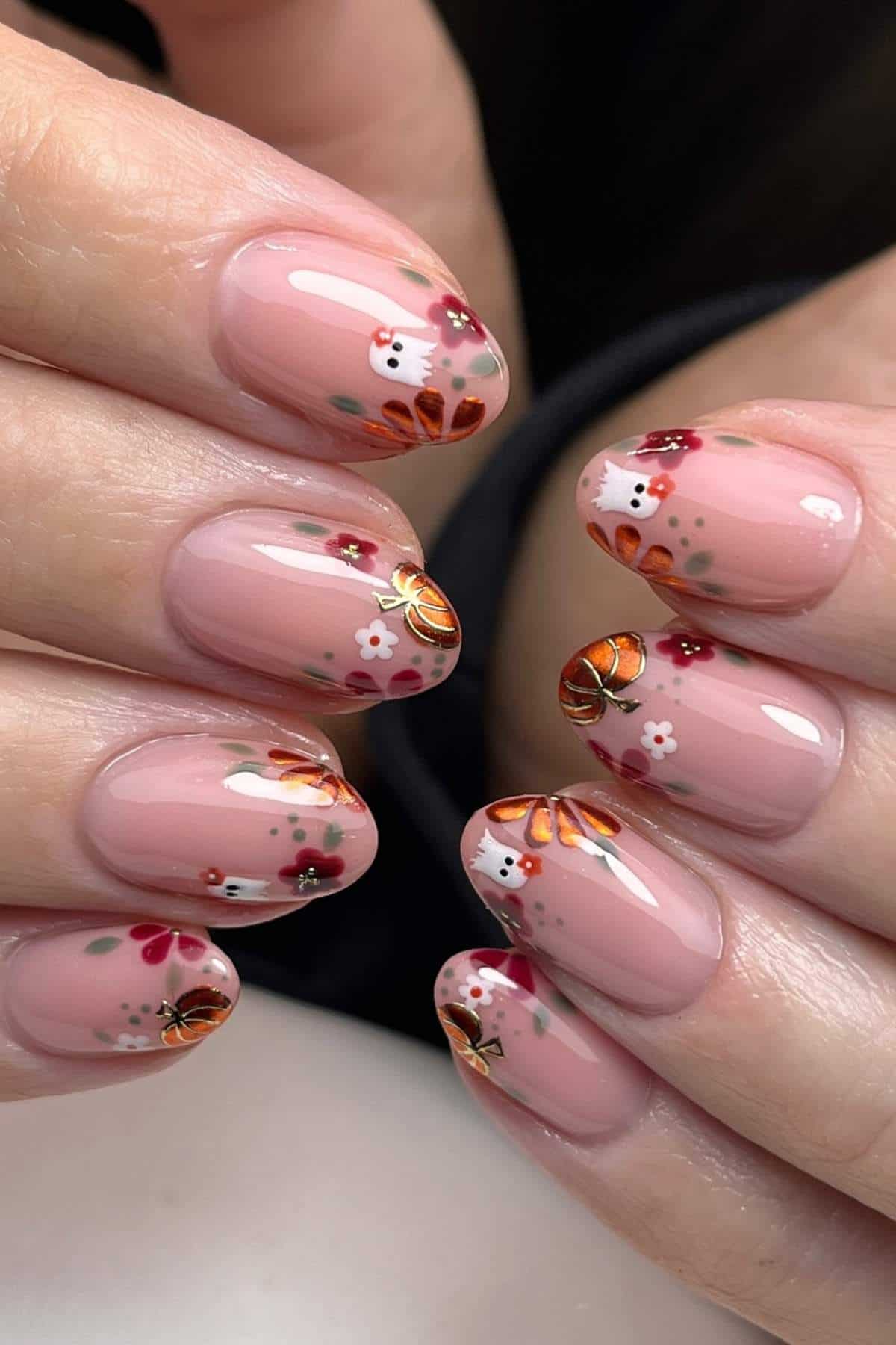 Hands with short, oval-shaped nails painted with a nude base and decorated with small red, white, and orange floral designs and metallic orange accents.