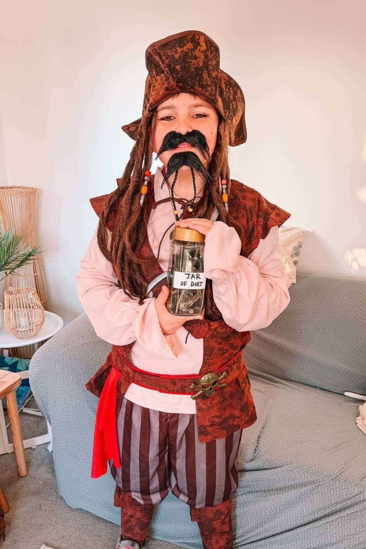 Person in a pirate costume holding a jar labeled “Jar of Dirt” stands indoors in front of a couch.