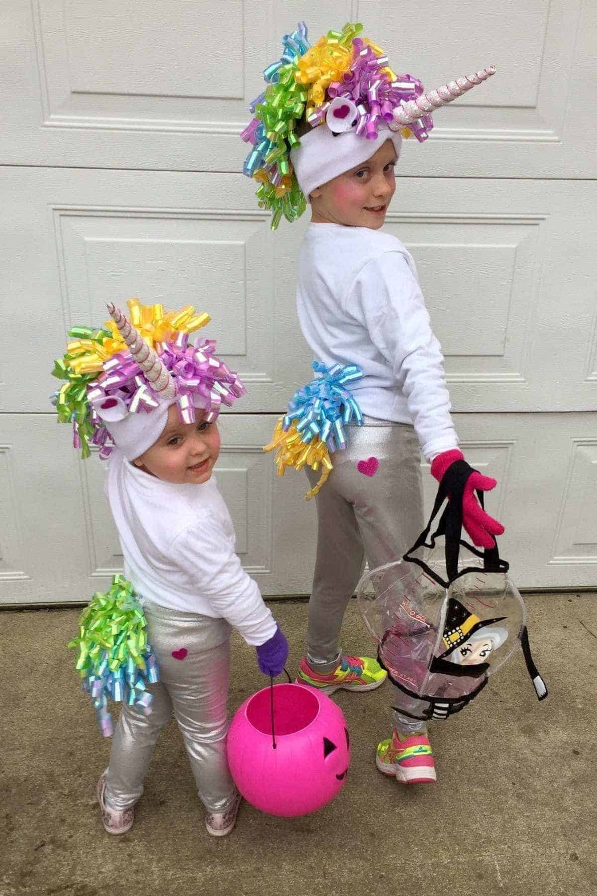 Two young children wear unicorn costumes with colorful bows, horns, and tails. One holds a pink pumpkin bucket, the other a clear bucket. They stand on a concrete surface in front of a garage door.