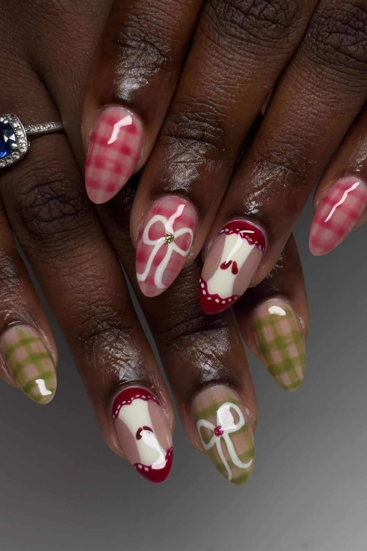 Hands with almond-shaped nails featuring red and green gingham patterns, bows, and heart designs on a neutral background. A ring with a blue stone is worn on one finger.