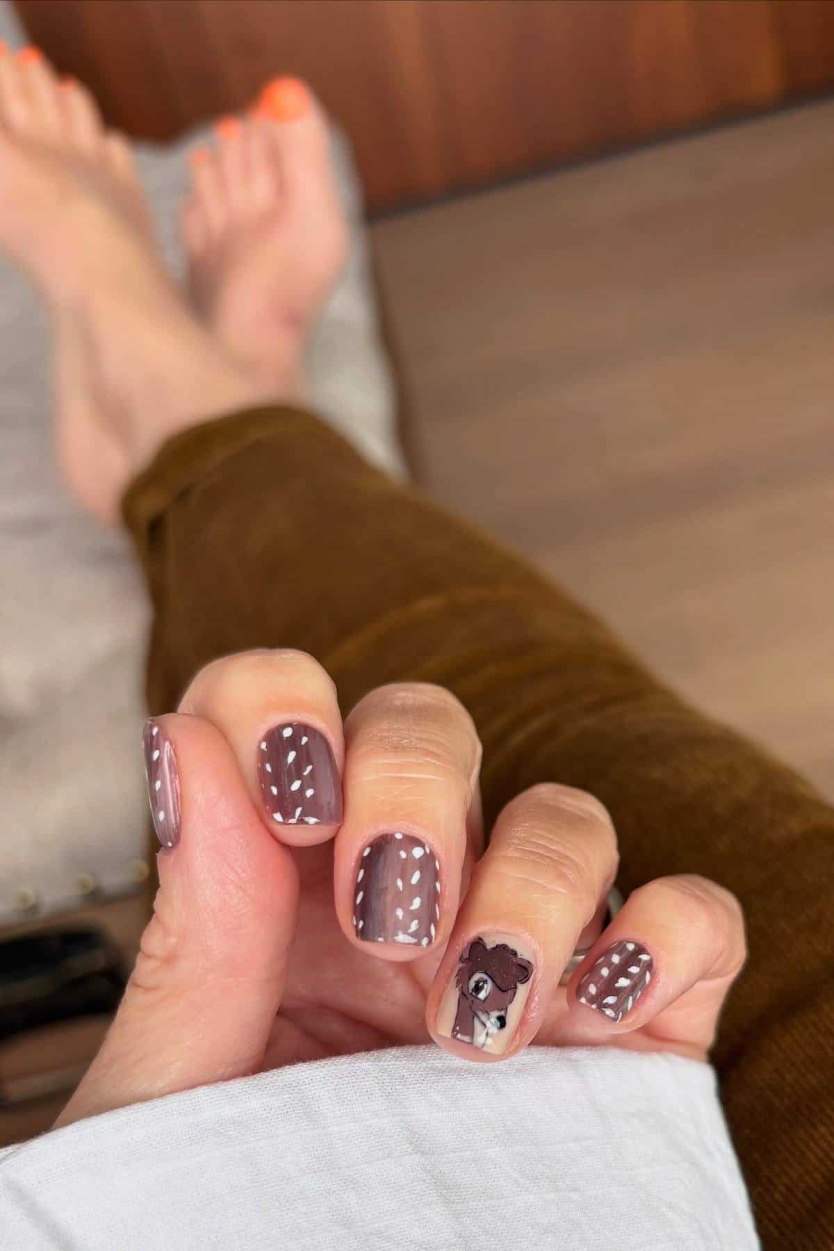 A hand with brown nail polish features white dotted lines and a deer face design on one nail; feet with orange toenails are visible in the background.