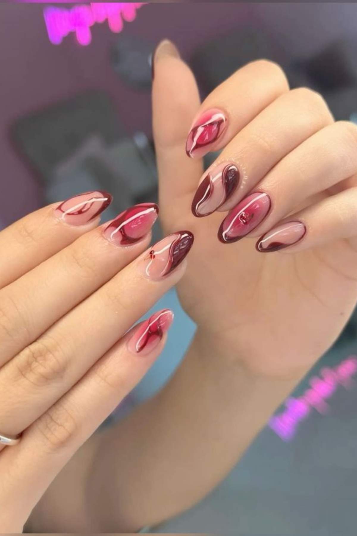 Close-up of hands with almond-shaped nails featuring glossy, abstract pink and burgundy nail art designs.