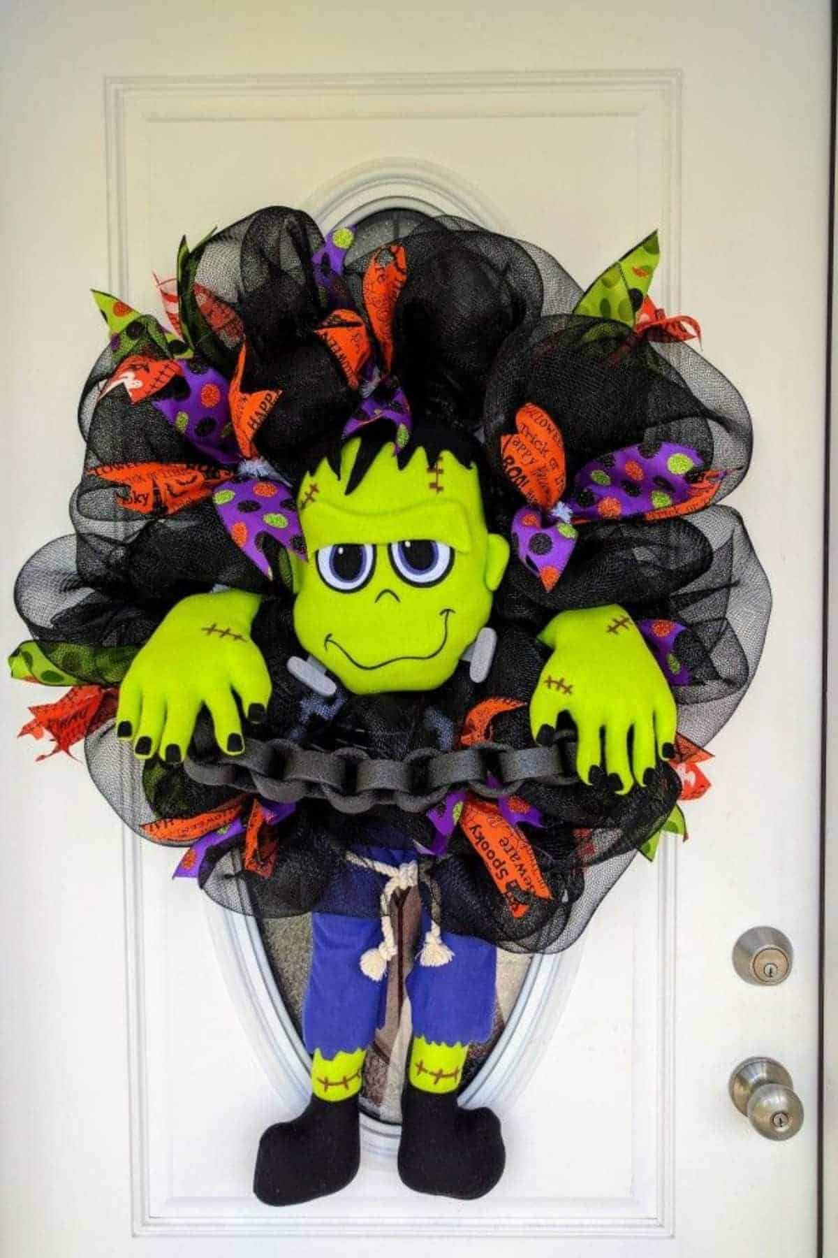 A Halloween wreath featuring a plush Frankenstein’s monster with a black chain, black mesh, and orange and purple ribbons hangs on a white door.