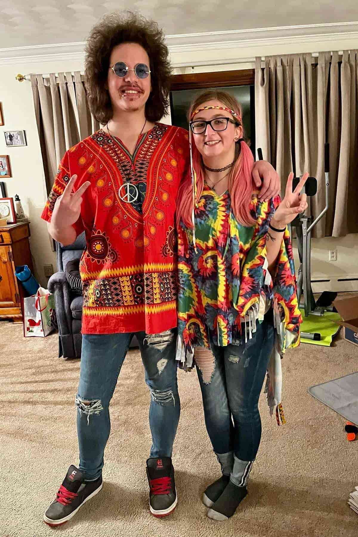 Two people dressed in 1960s-style hippie outfits, both making peace signs and smiling indoors in a living room.
