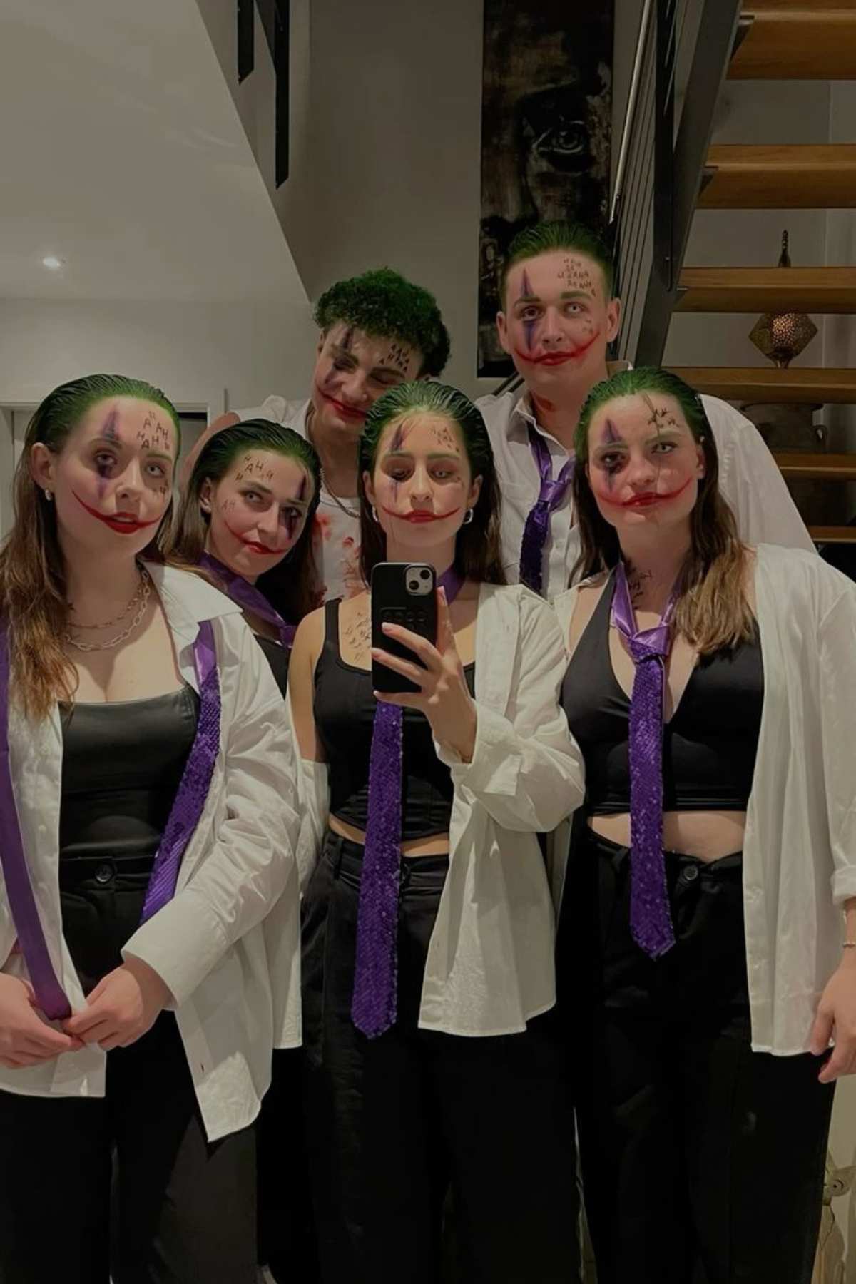 Five people dressed in black pants, white shirts, purple ties, and Joker-inspired makeup pose for a group selfie indoors.