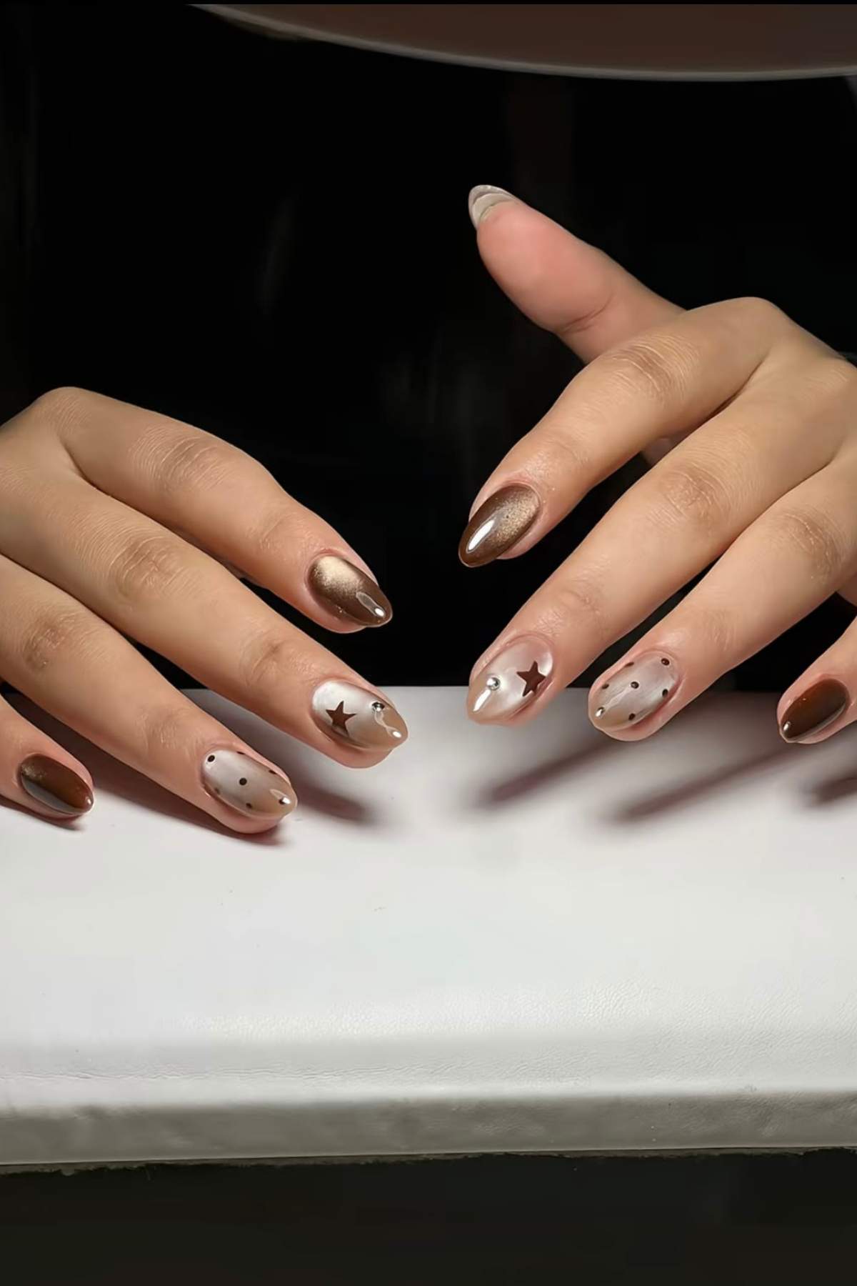 Hands with almond-shaped nails featuring metallic bronze polish and accent nails with beige polish, black dots, and star designs, resting on a white surface.