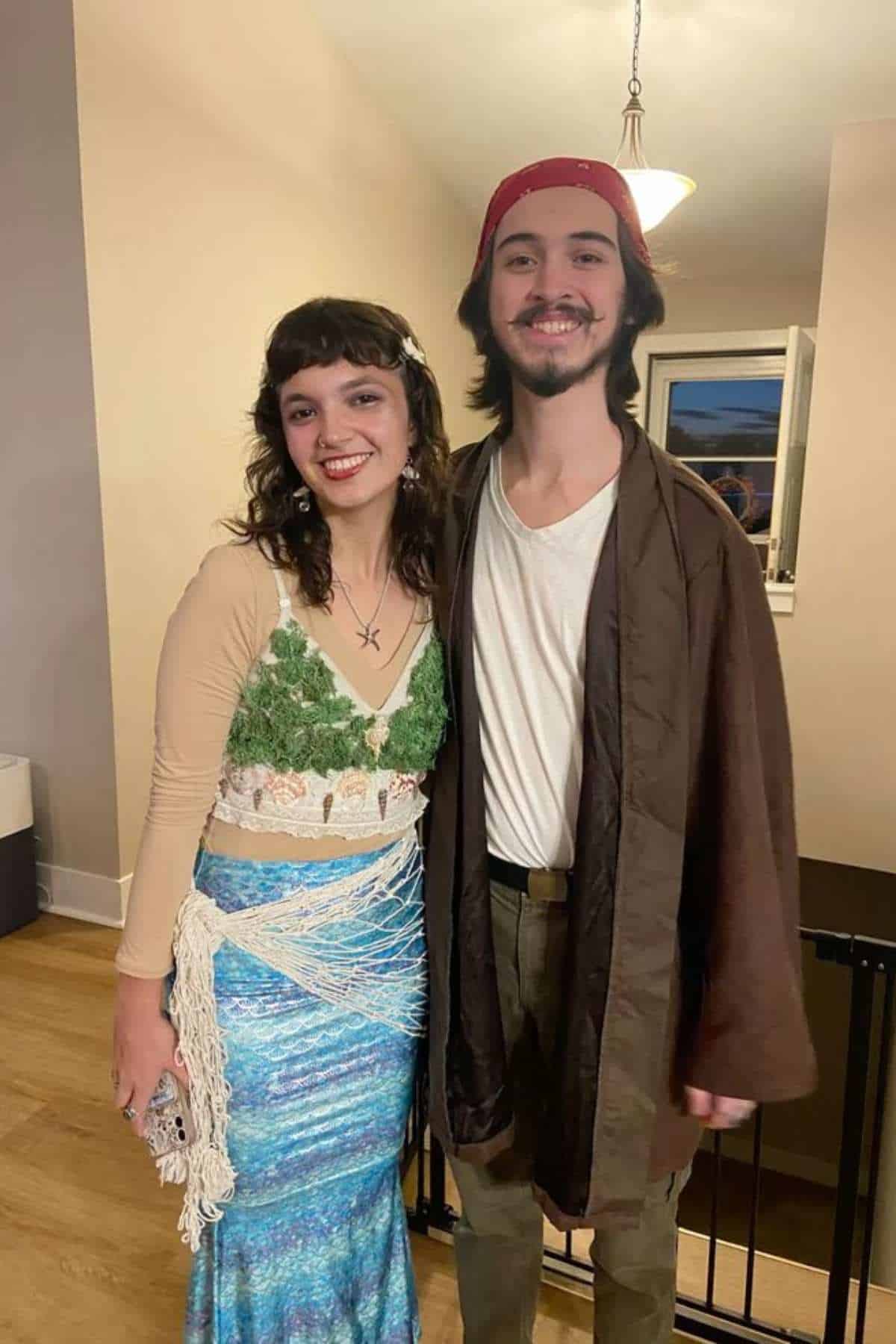 Two people in costume stand indoors; the woman wears a nature-themed outfit and the man wears a brown robe, white shirt, red headscarf, and belt. Both are smiling at the camera.