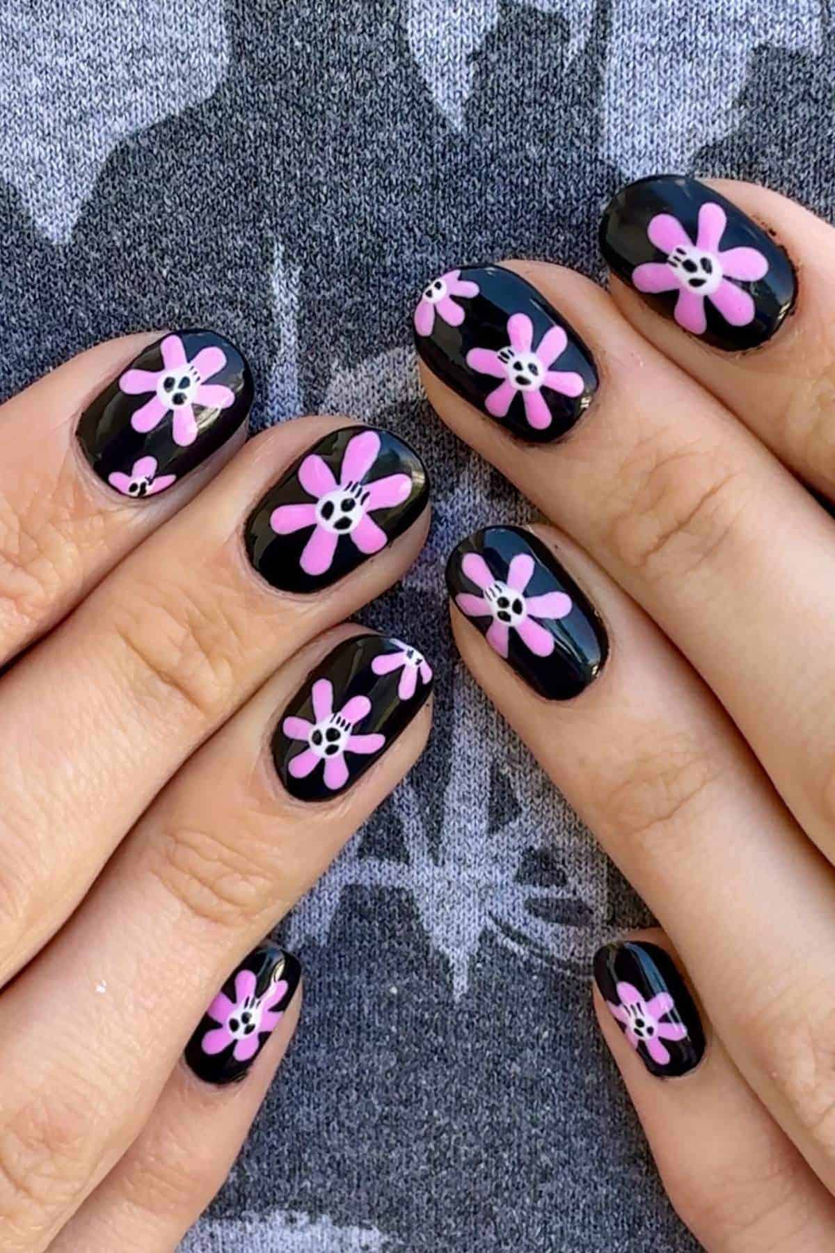 Hands with black nail polish and pink and white flower designs on each nail, shown against a gray patterned fabric background.