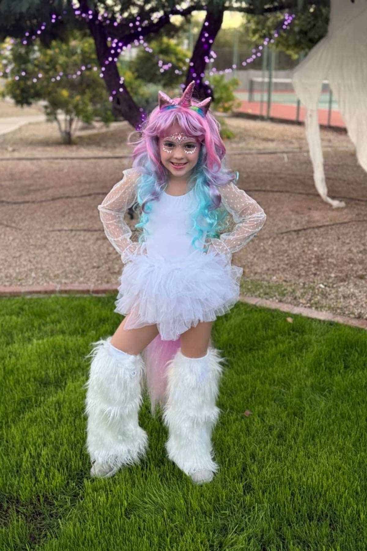 Child in a unicorn costume with a pastel wig, unicorn horn, white tutu dress, and furry leg warmers, standing on grass outdoors.
