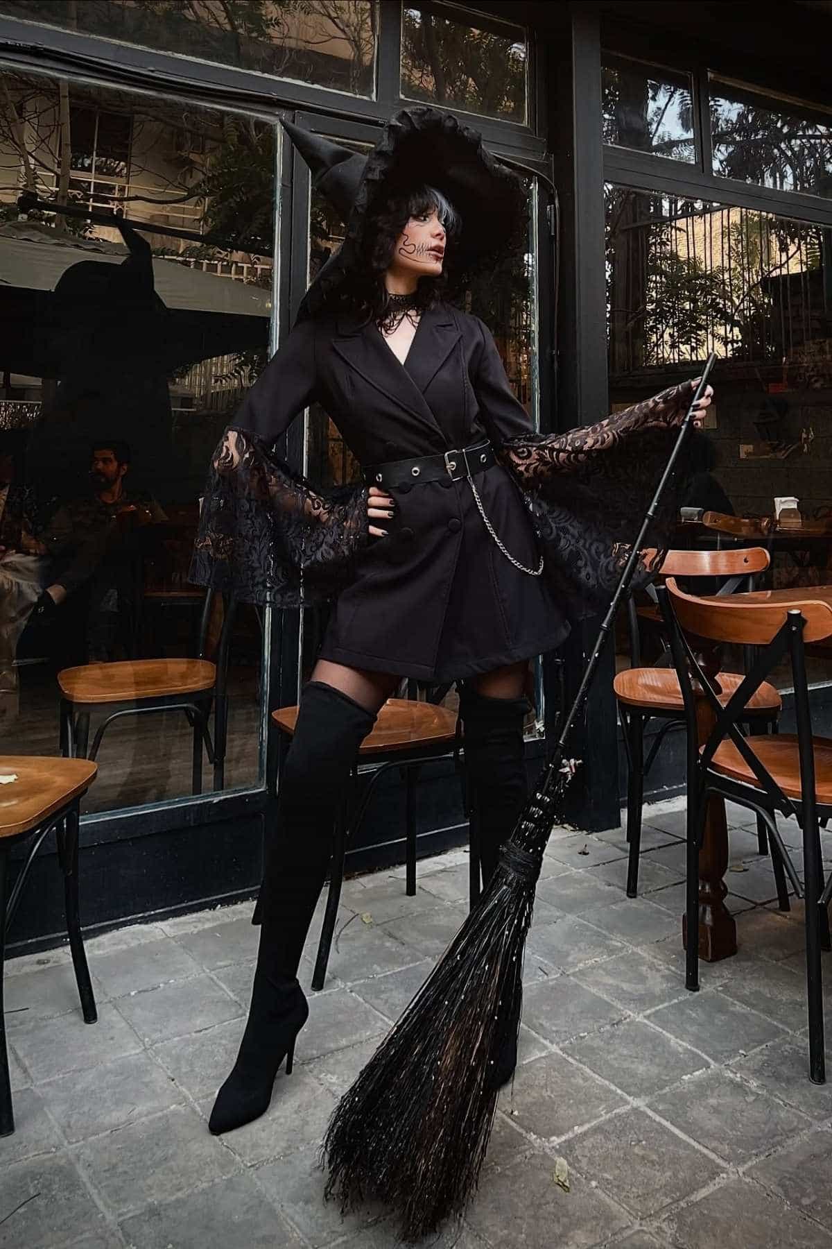Person wearing a black witch costume with a pointed hat and lace sleeves stands outdoors holding a broom, surrounded by wooden chairs and tables.