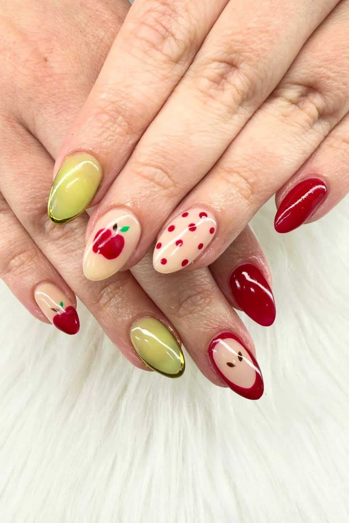 Hands with almond-shaped nails featuring nail art: red apples, green polish, red tips, dots, and minimalistic fruit designs on a white surface.