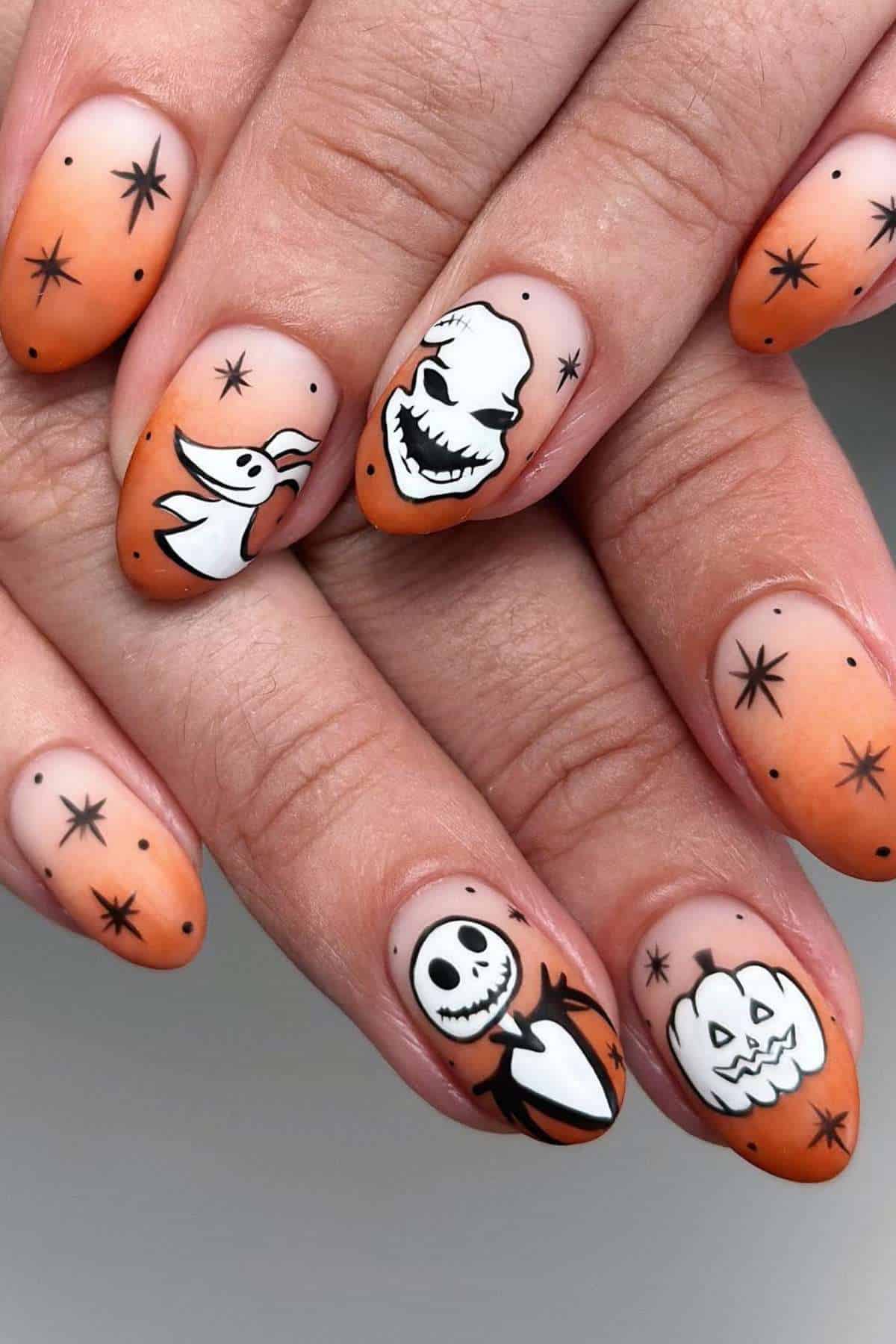 Fingernails painted with an orange gradient and black stars, each nail featuring Halloween-themed cartoon faces in black and white.