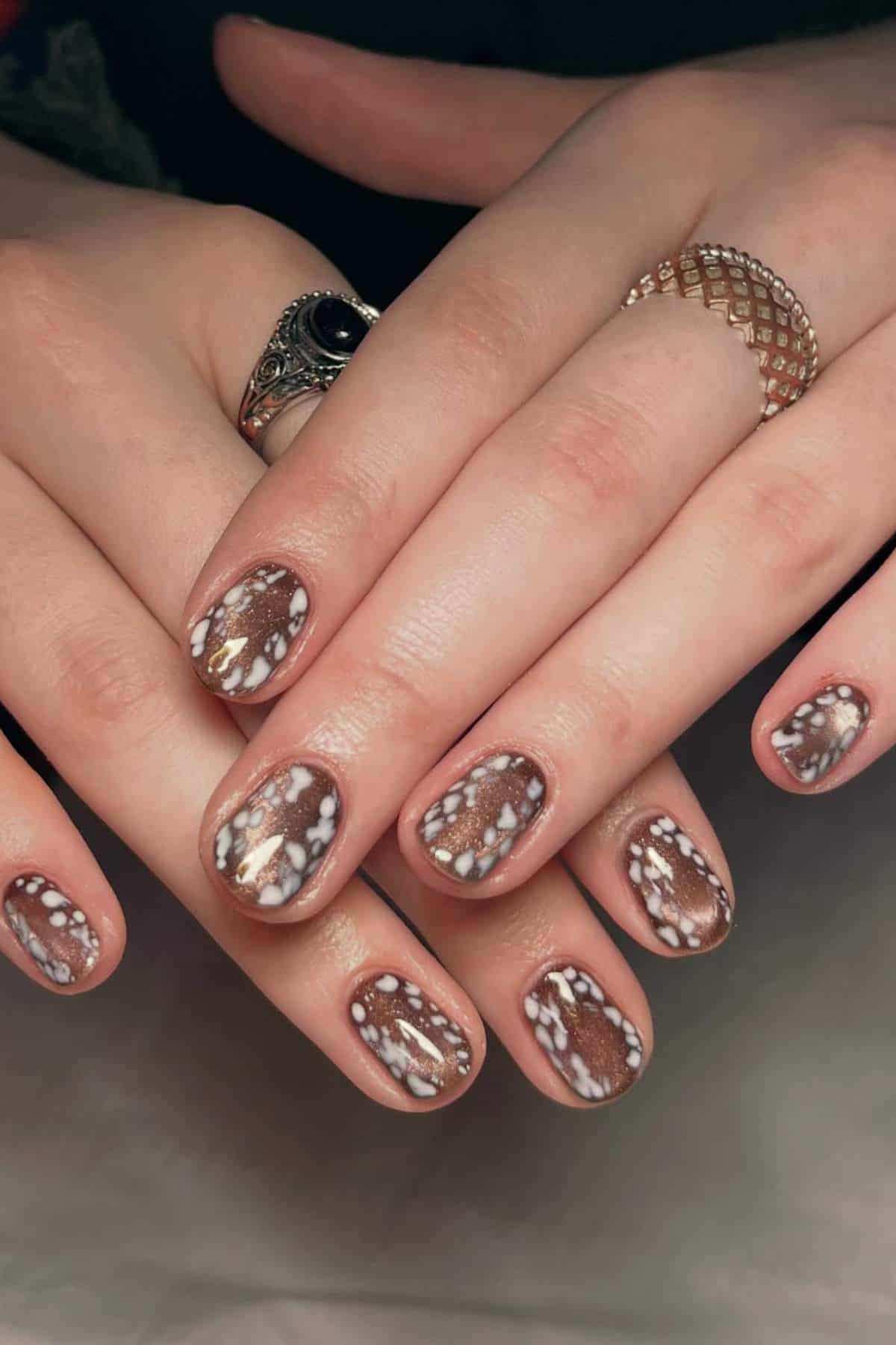 Hands with short, oval nails feature a brown base color and white, speckled patterns resembling deer print. The person wears two rings, one with a black stone and another textured band.