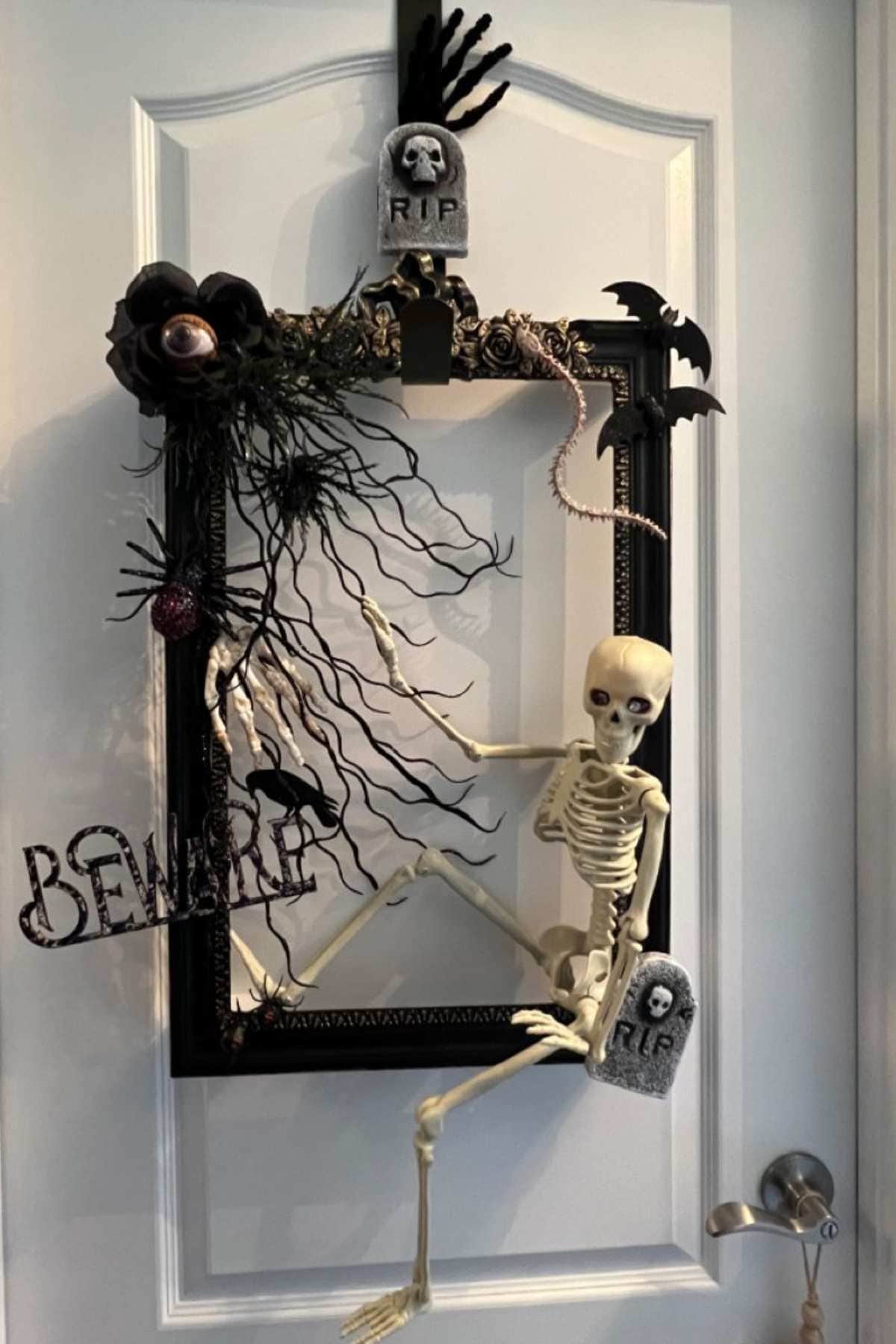 A Halloween decoration featuring a skeleton, tombstones, fake plants, a bat, and a "Beware" sign arranged in a black picture frame on a white door.