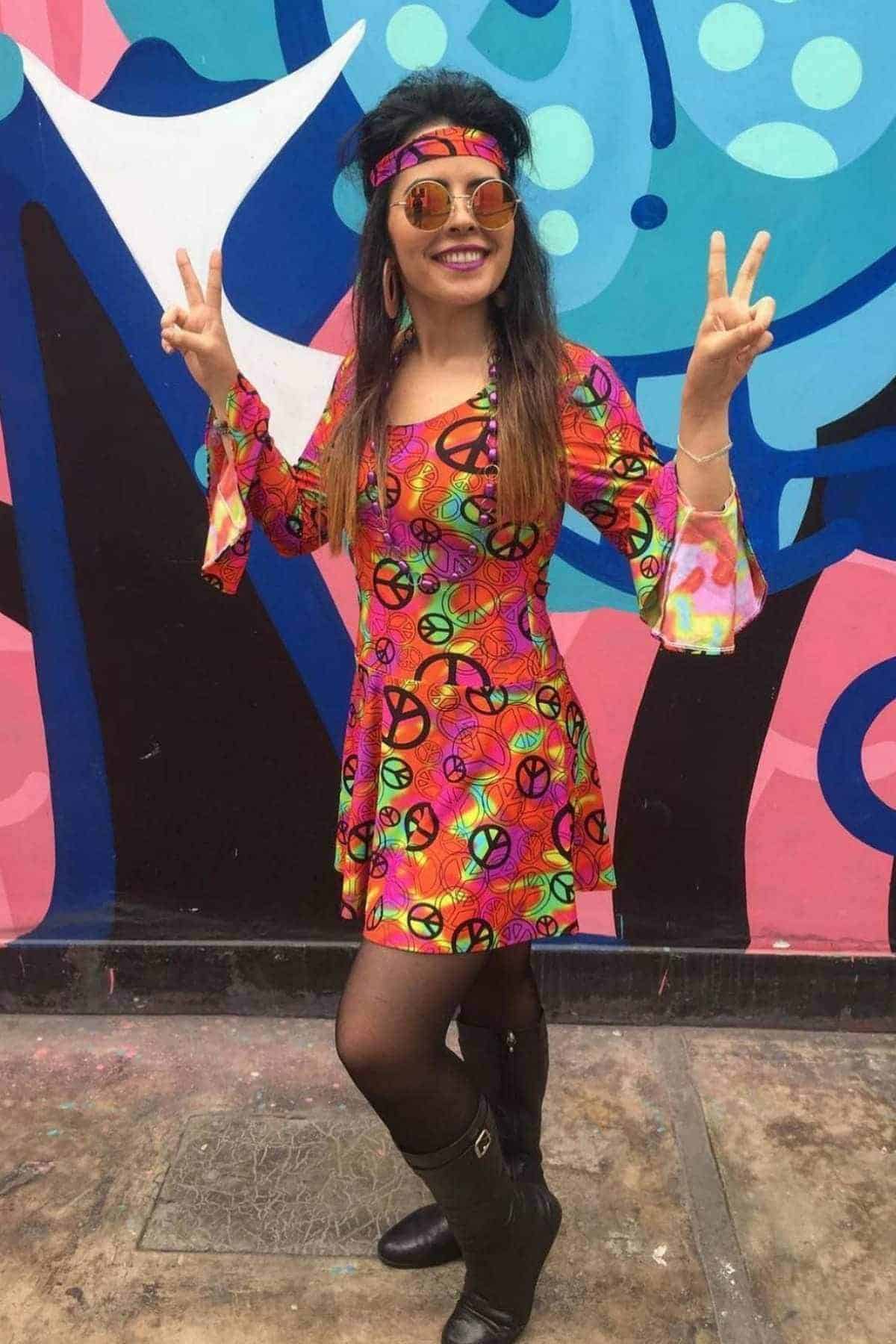 Woman in a colorful 1970s-style dress with peace symbols, headband, sunglasses, and knee-high boots poses in front of a vibrant mural, making peace signs with both hands.
