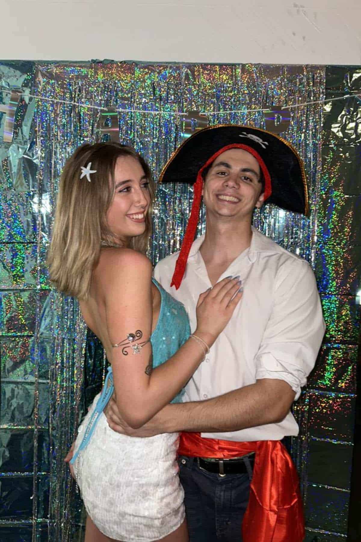 Two people pose together at a party; one wears a pirate hat and sash, the other has butterfly wings and a star hair clip, with a shiny, reflective backdrop behind them.