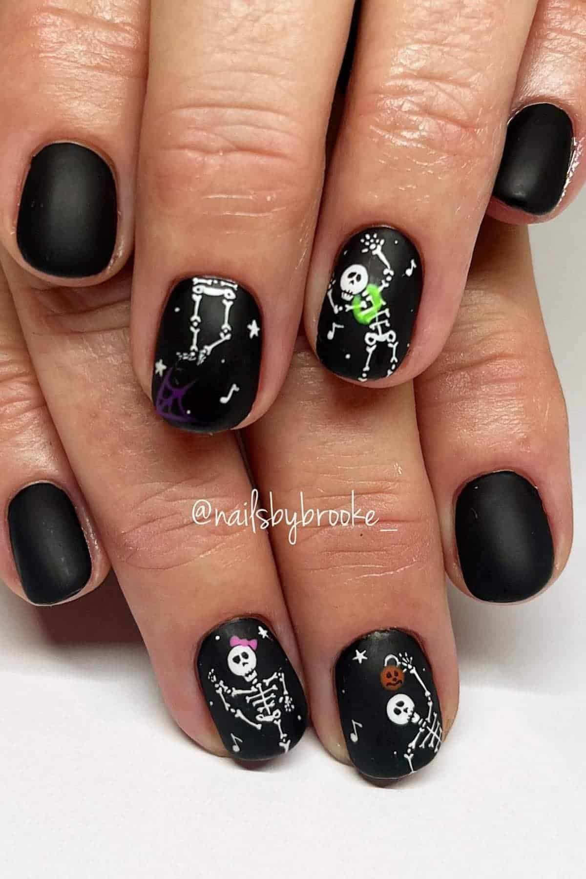 Black matte nails with white skeleton designs, stars, and colorful Halloween details on two accent nails.