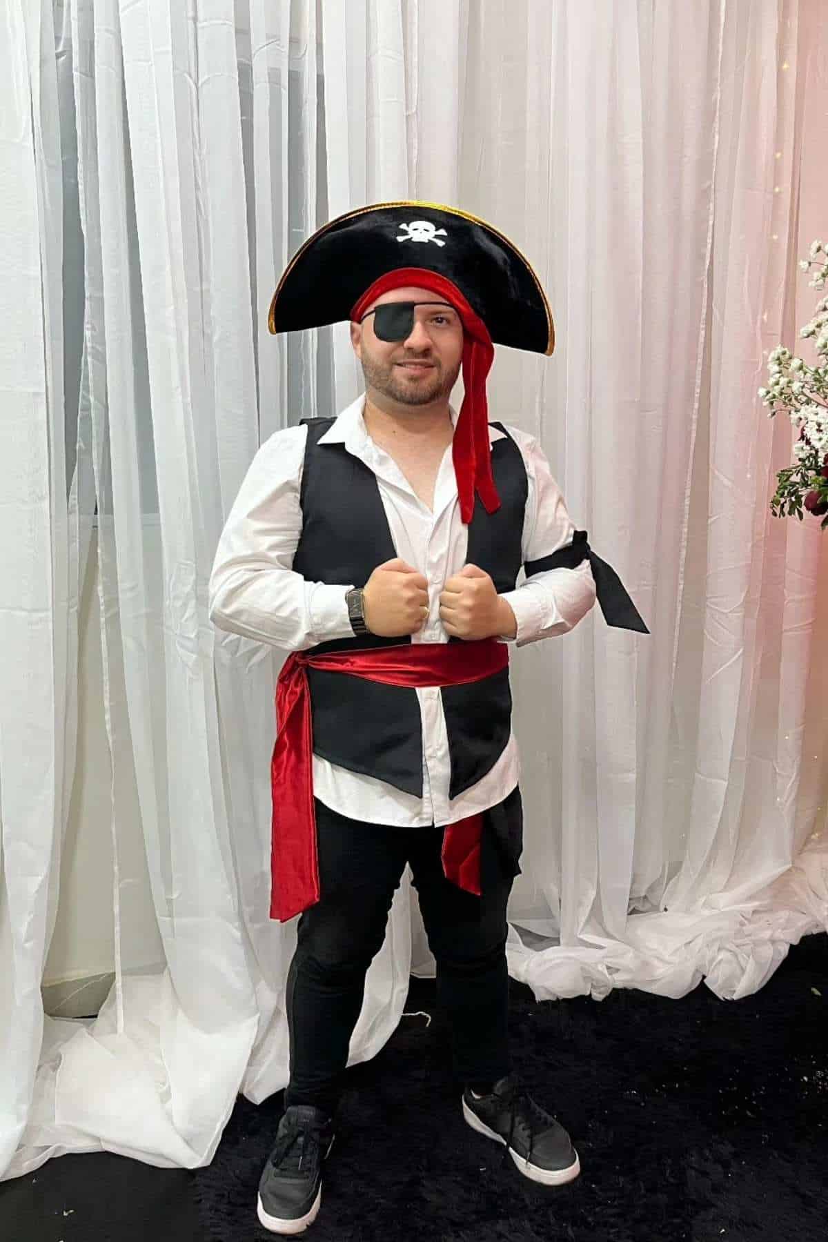 A person dressed in a pirate costume with a hat, eye patch, red sash, and black boots stands in front of white drapes and flowers.