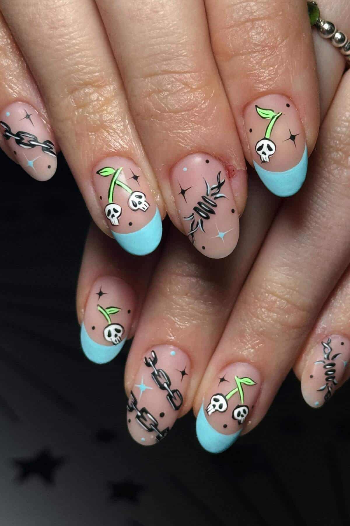 Almond-shaped nails with light blue tips and nail art featuring skulls, green sprouts, chains, barbed wire, and small stars on a natural base.