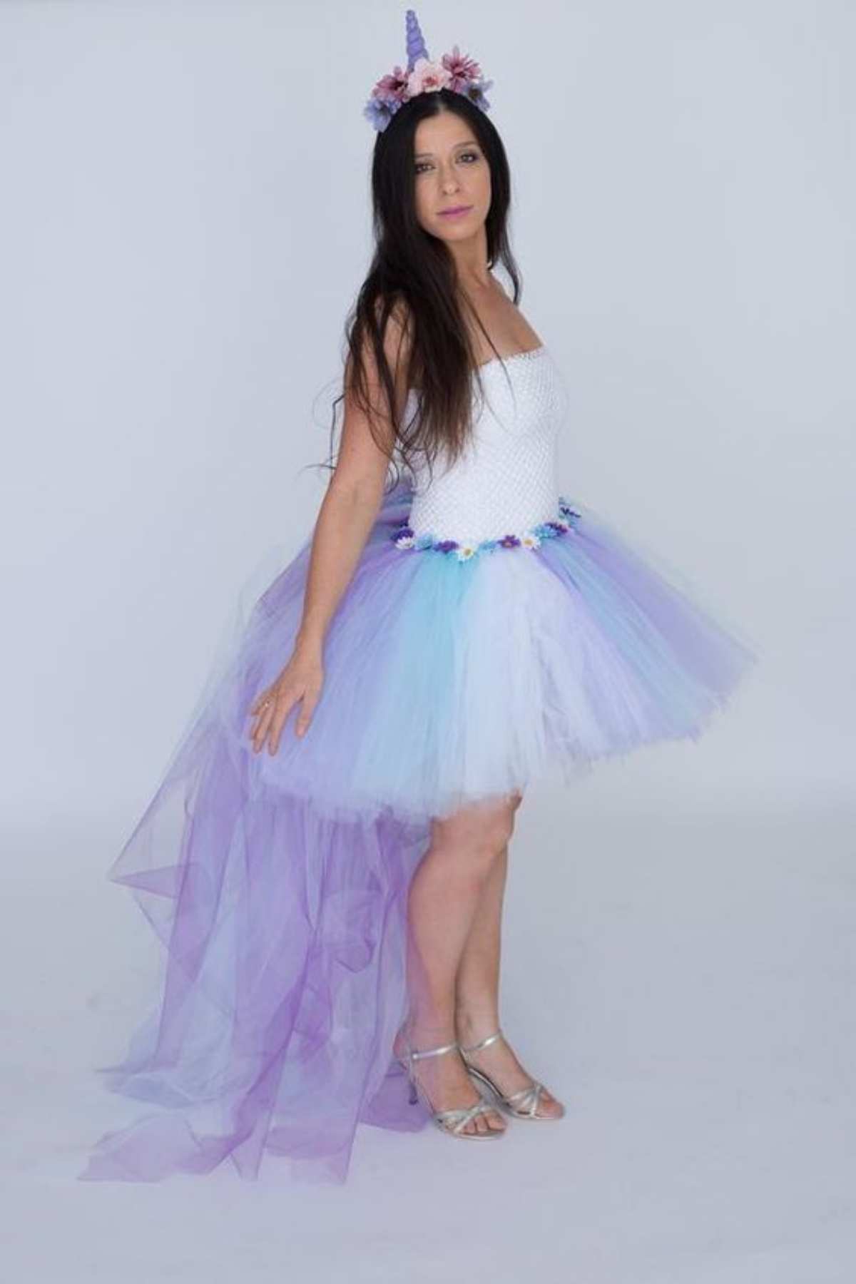 Woman wearing a white top, tulle skirt with a long purple train, and a unicorn horn headband with flowers, standing against a plain background.