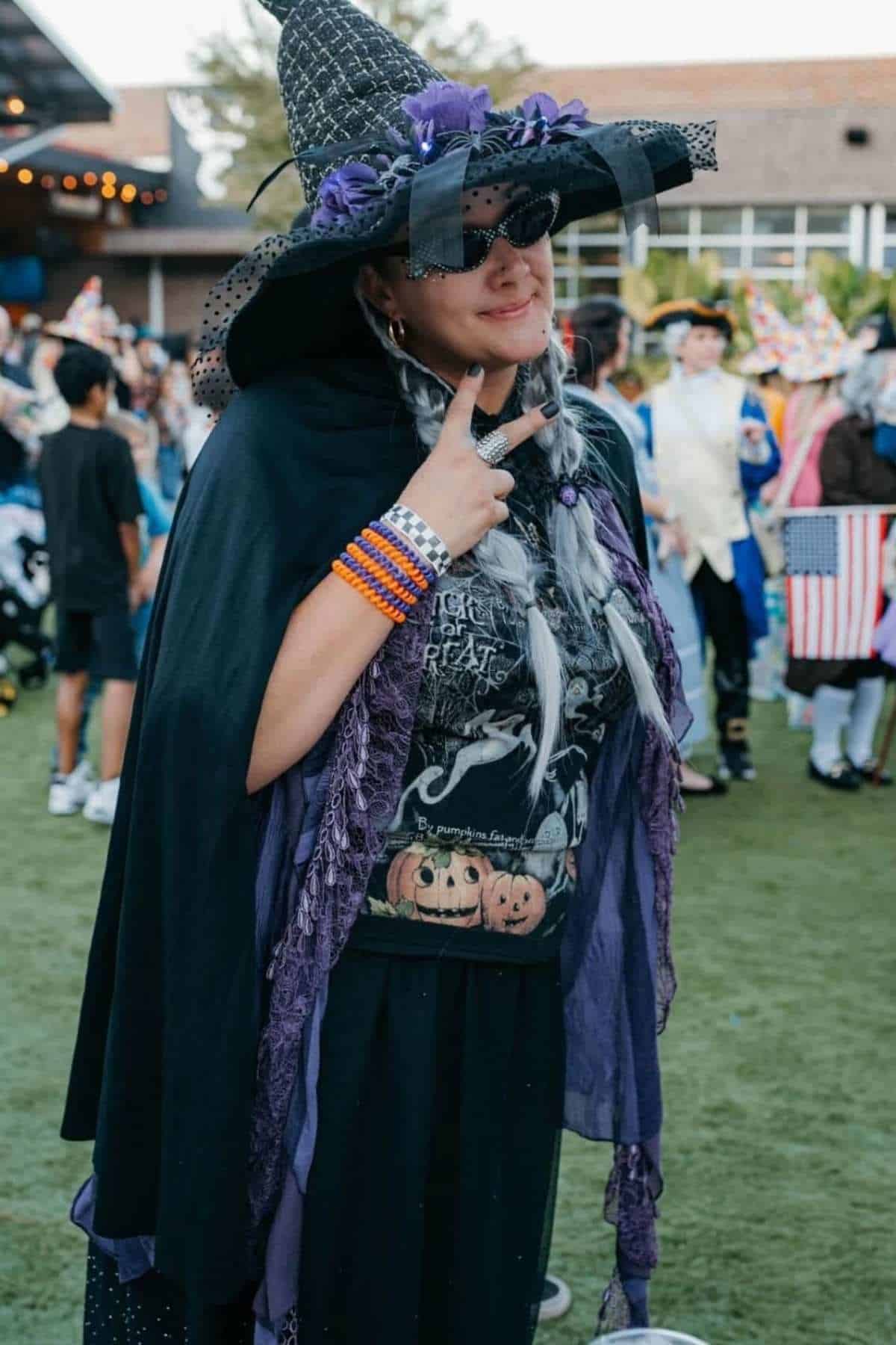 Person wearing a witch costume with a pointed hat, black cape, and Halloween-themed shirt, making a peace sign at an outdoor event.