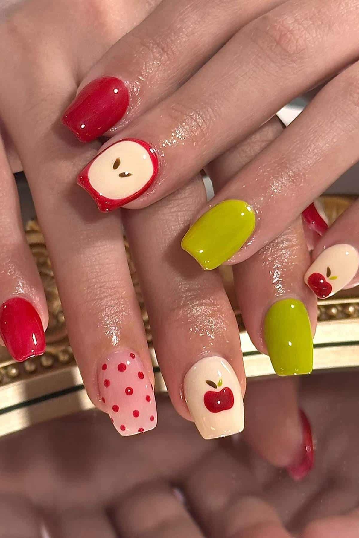 Close-up of hands with colorful nail art featuring red, green, and beige polish, with designs of apples, apple slices, and red dots on some nails.