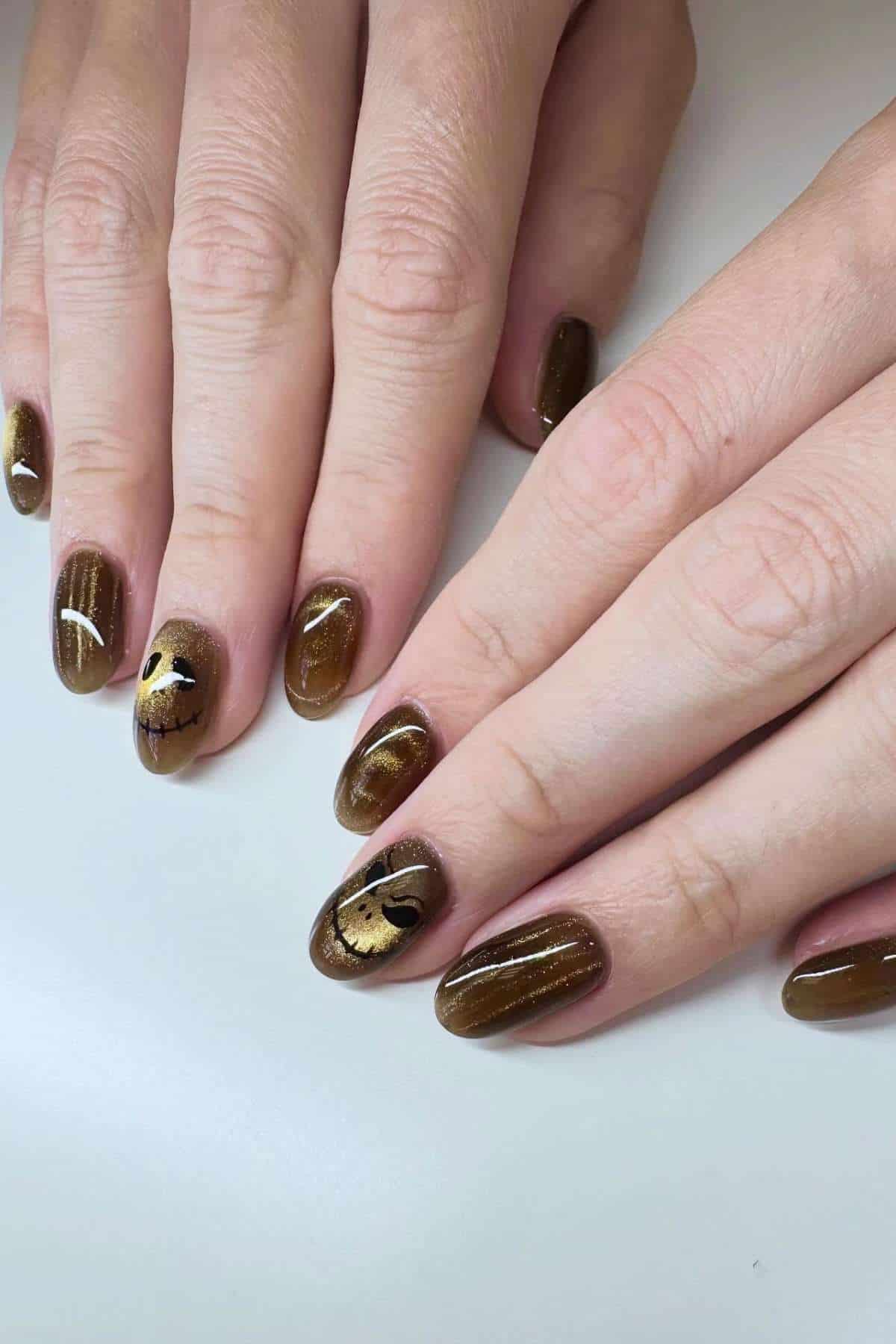 Hands with brown, glossy oval-shaped nails; some nails feature gold foil and black line art accents.