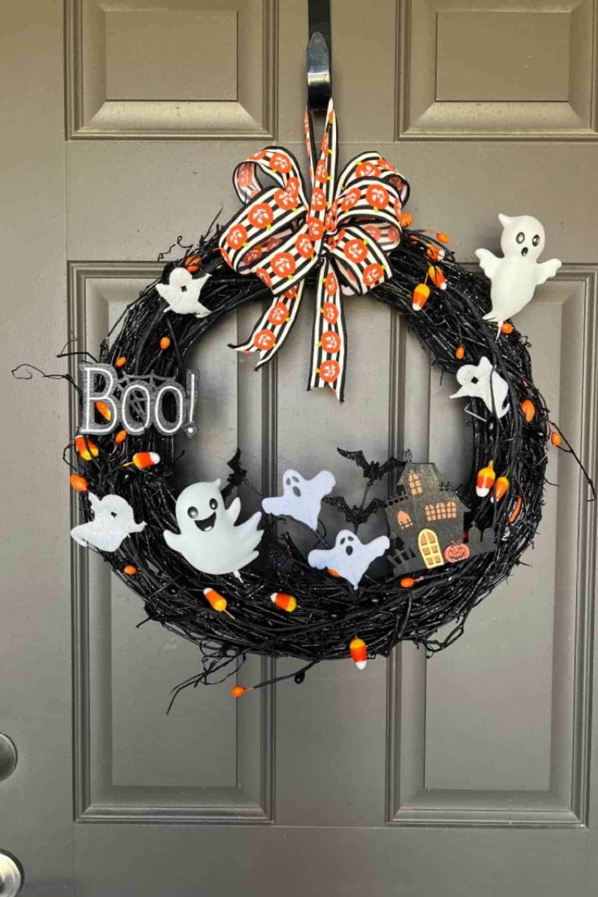 Black wreath with ghosts, a haunted house, candy corn, "Boo!" sign, and a bow with pumpkin pattern hanging on a gray door.