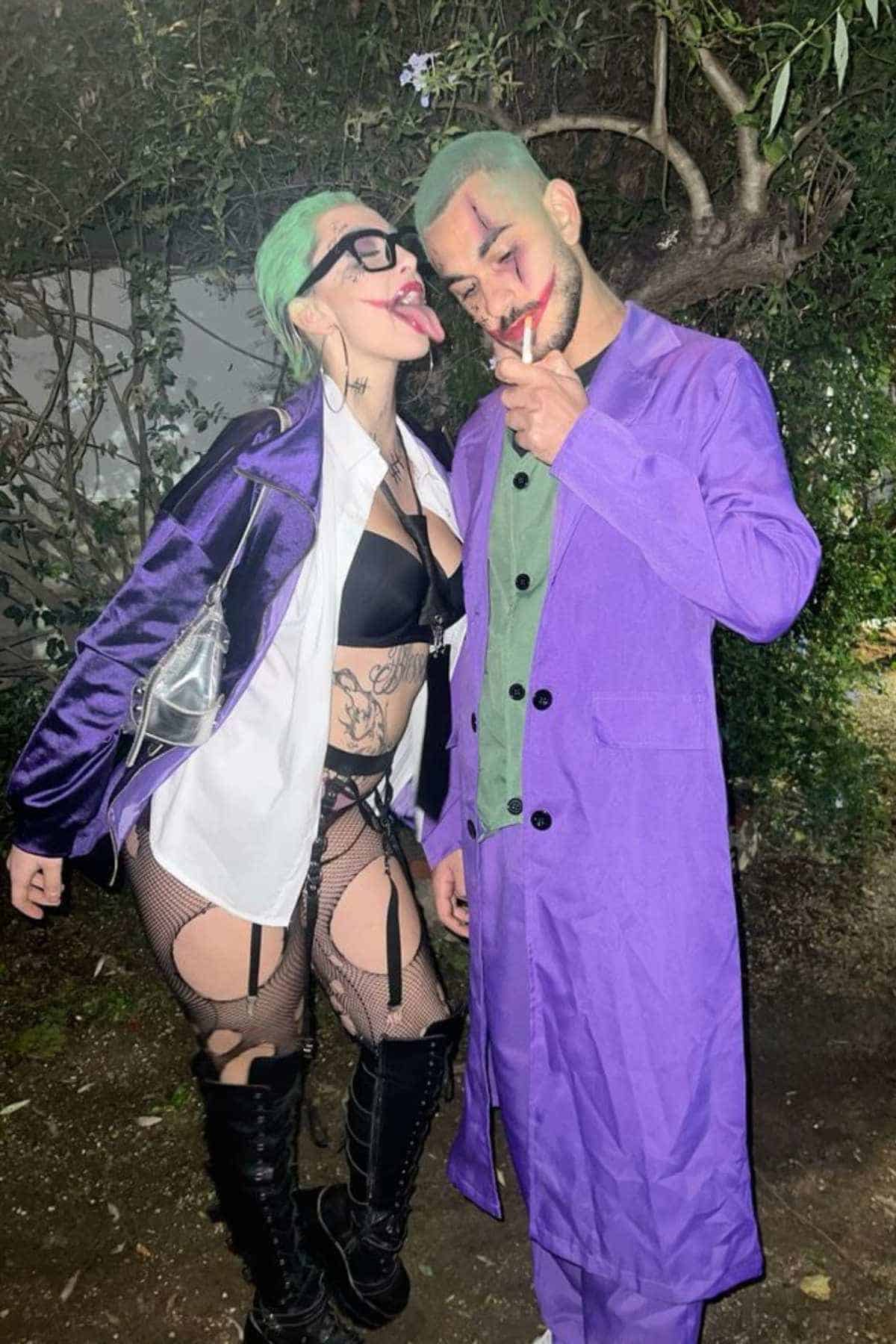 Two people dressed in Joker-inspired costumes pose outdoors at night; one person playfully sticks out their tongue, both wear purple coats and have green hair.