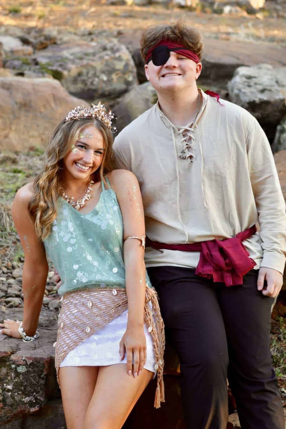 Two people in costumes pose outdoors; one wears a tiara and sparkly outfit, the other wears an eye patch, tunic, and red sash.