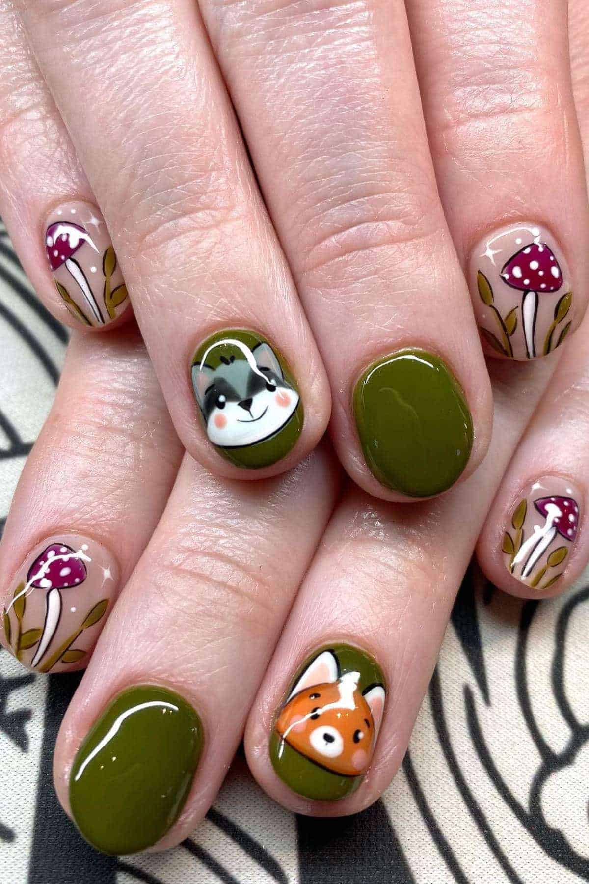Hands with short nails painted olive green and nude; designs include a cartoon fox, a rabbit, and small red mushrooms with white spots on some nails.