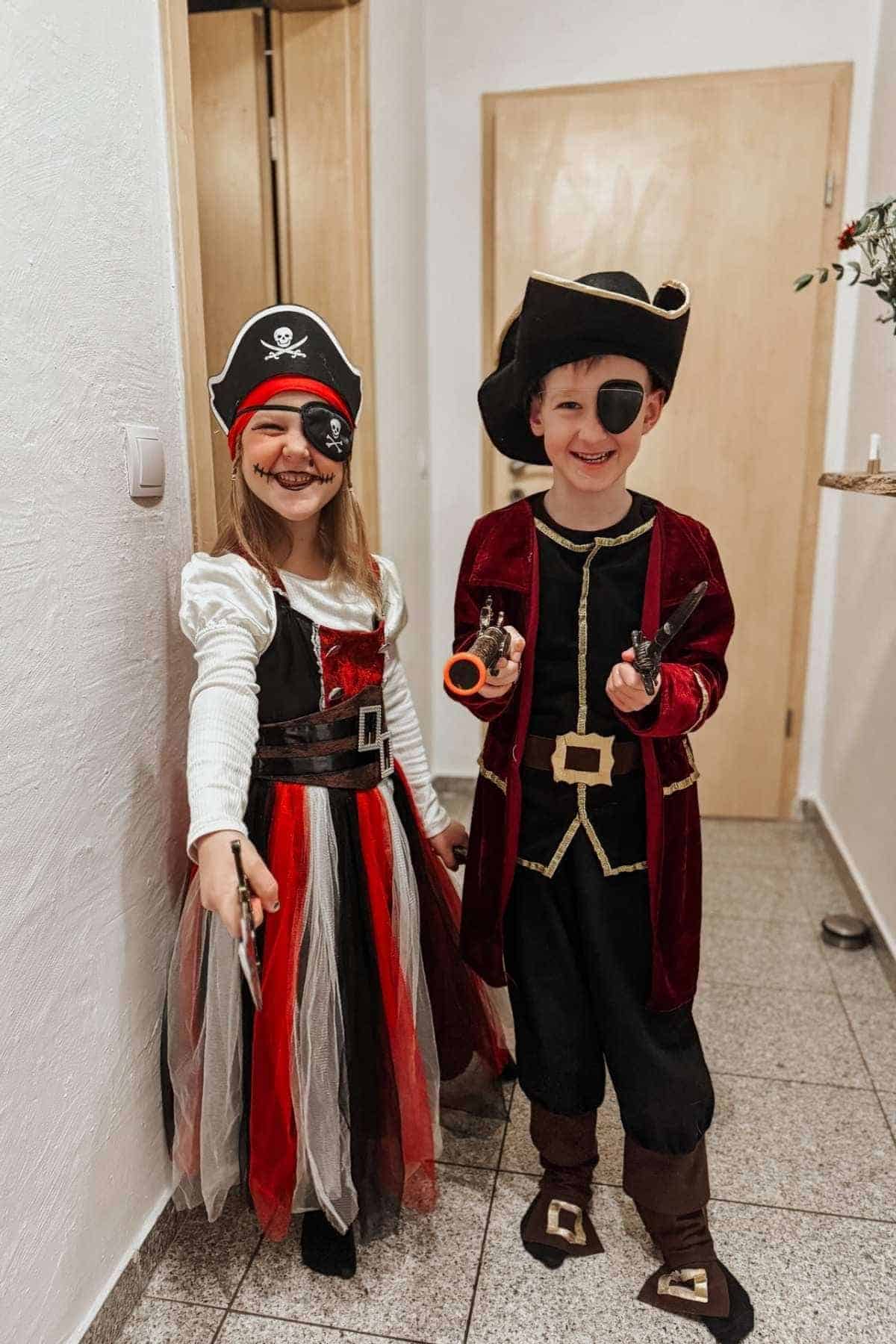 Two children dressed as pirates stand in a hallway, both smiling and wearing eye patches, hats, and holding toy weapons.