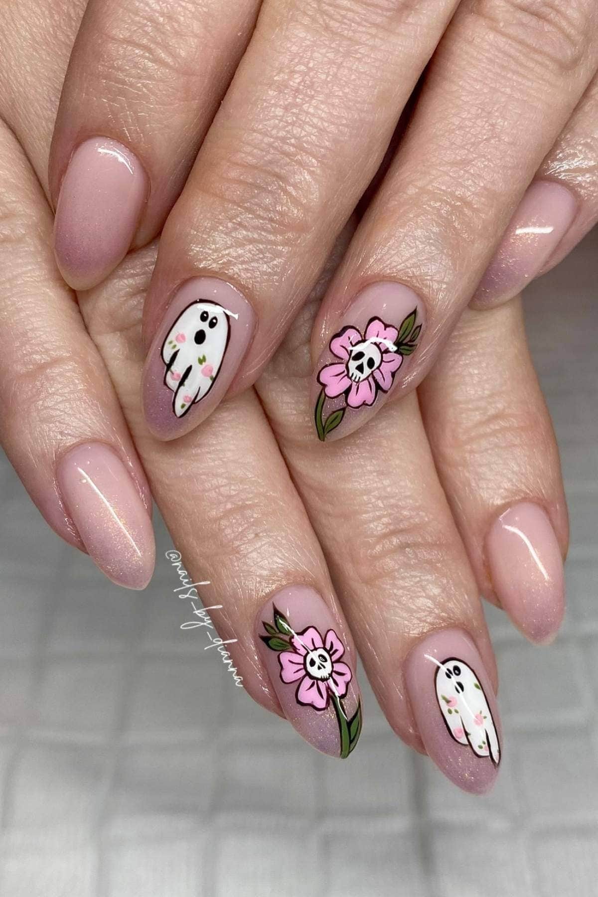 Hands with almond-shaped nails featuring a light pink ombre polish, two nails with ghost and pink flower designs, and the rest in plain ombre.