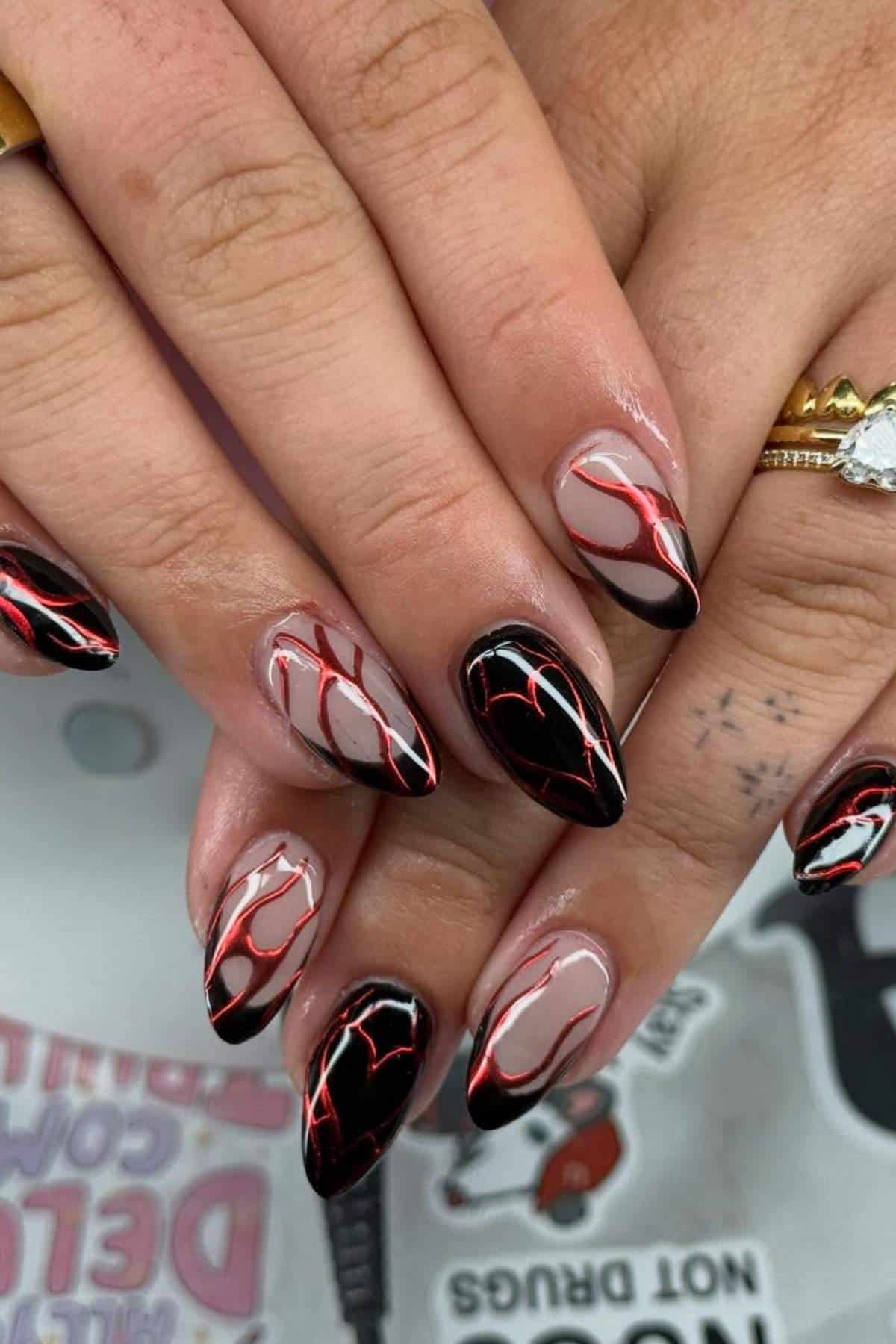 Hands with almond-shaped nails featuring a nude base and black, red, and white flame-like nail art. Some fingers have visible tattoos and gold rings.