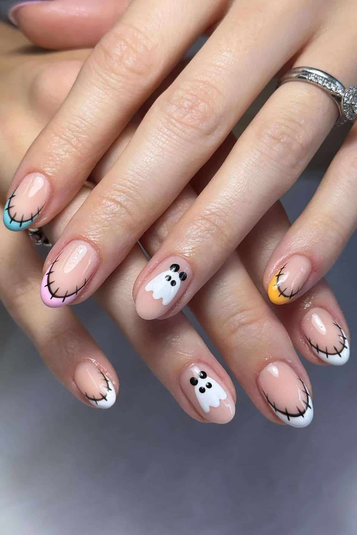 Hands with Halloween-themed nail art featuring stitched tips and small ghost designs on a nude base.