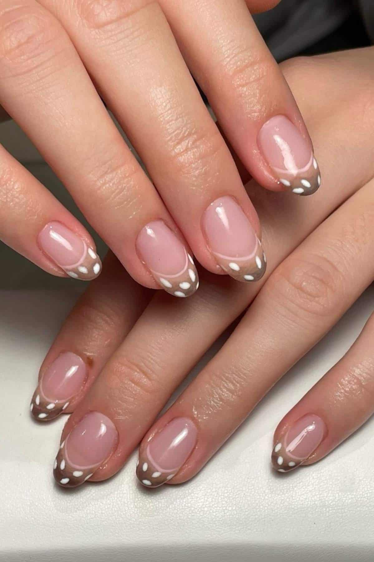 Hands with manicured nails featuring a nude base, brown French tips, and small white dot accents on each tip.