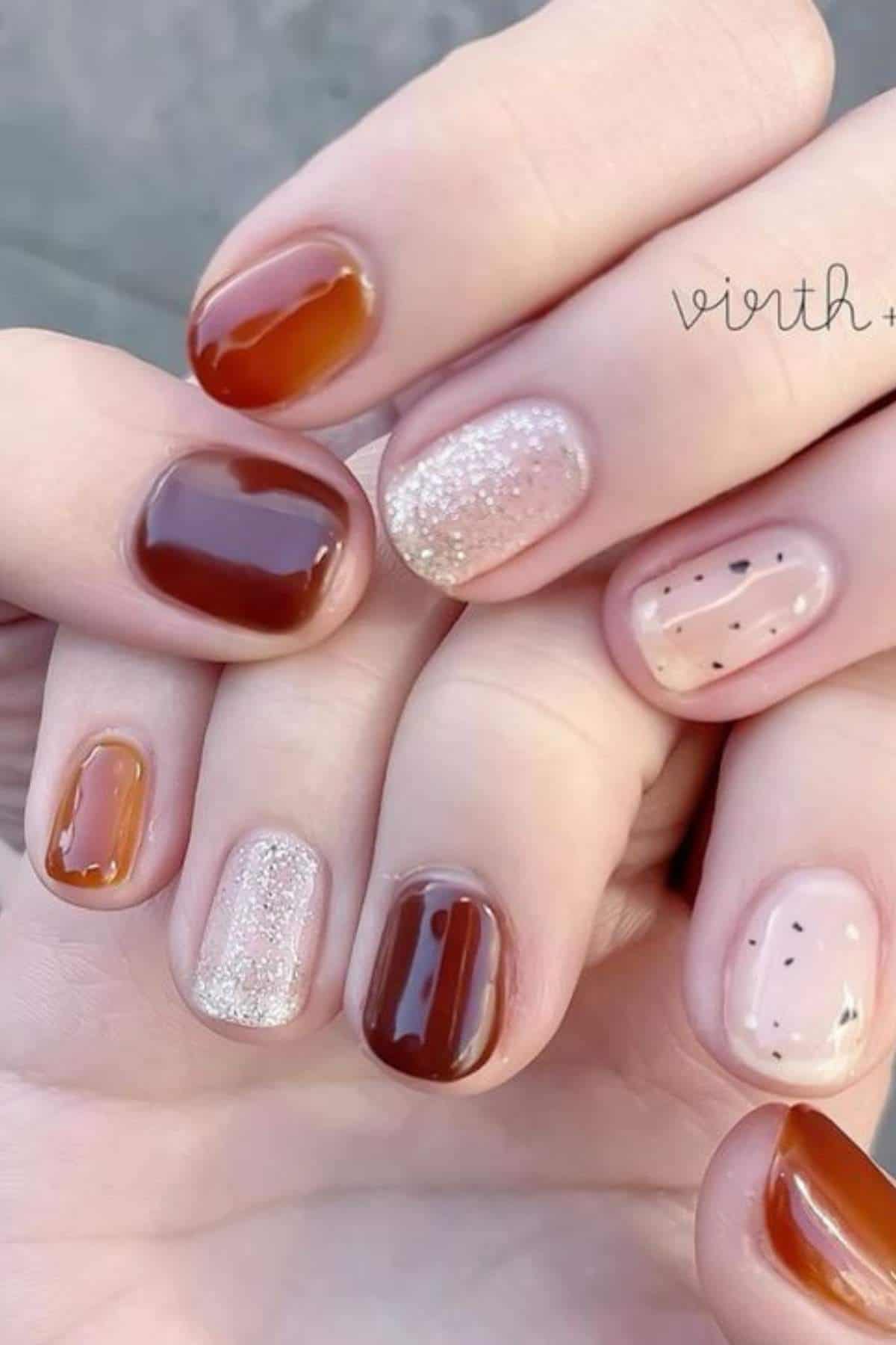 Close-up of hands with short nails painted in shades of brown, nude, glitter, and speckled beige, featuring a glossy finish.