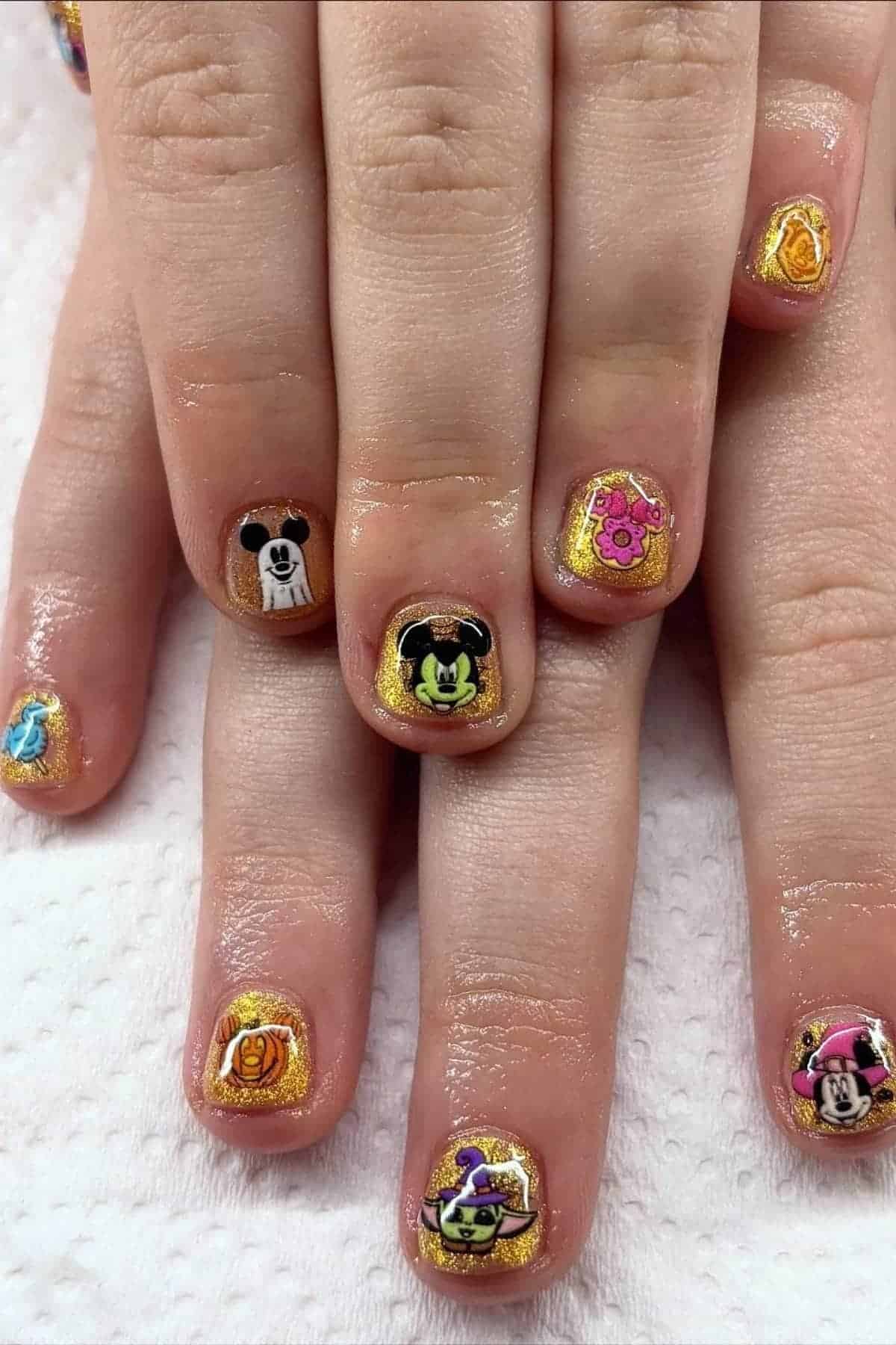 Hands with short fingernails painted gold and decorated with colorful cartoon character stickers, including various animals and faces, resting on a white textured surface.