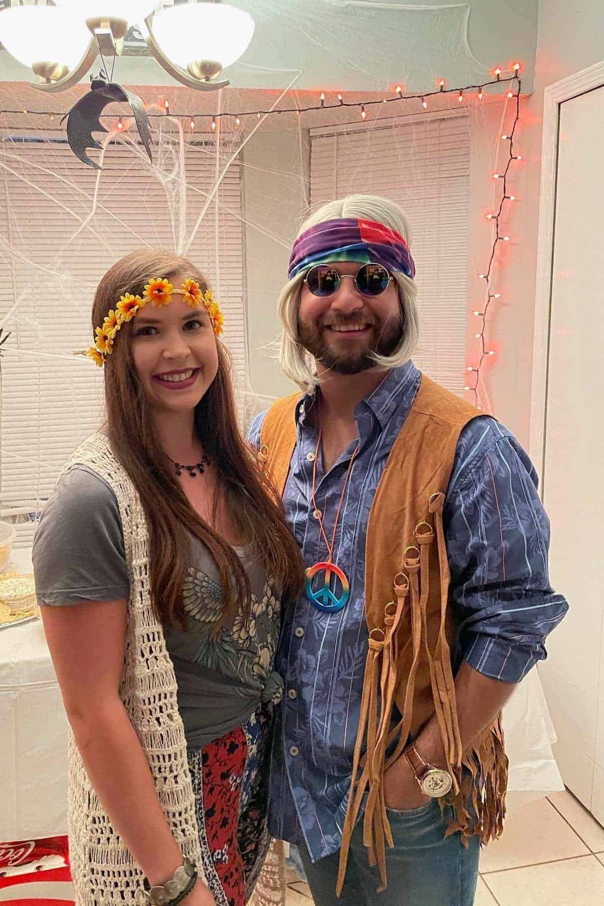 Two people pose indoors wearing 1970s-inspired costumes; the woman has a flower headband and vest, the man wears a headband, sunglasses, fringed vest, peace necklace, and wig.