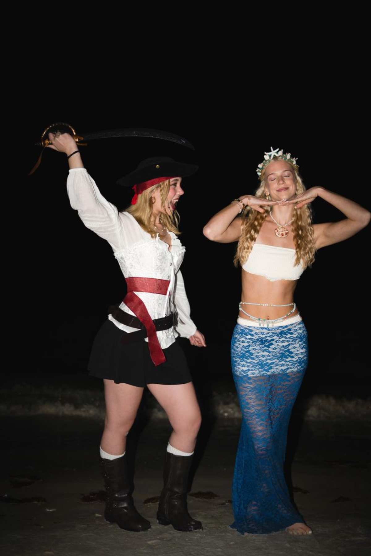 Two women in costumes stand on a dark beach; one dressed as a pirate holding a sword, the other as a mermaid posing with hands under her chin.
