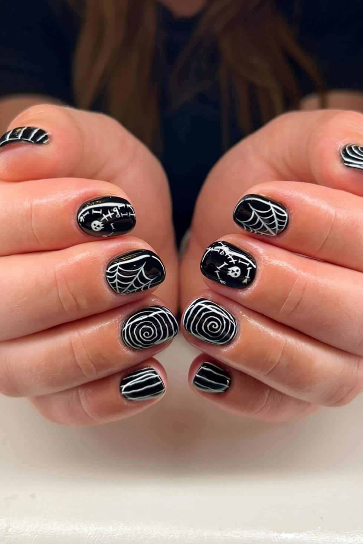 Hands with short black nails featuring white Halloween-themed designs, including spiderwebs, spirals, stripes, and graveyard symbols.