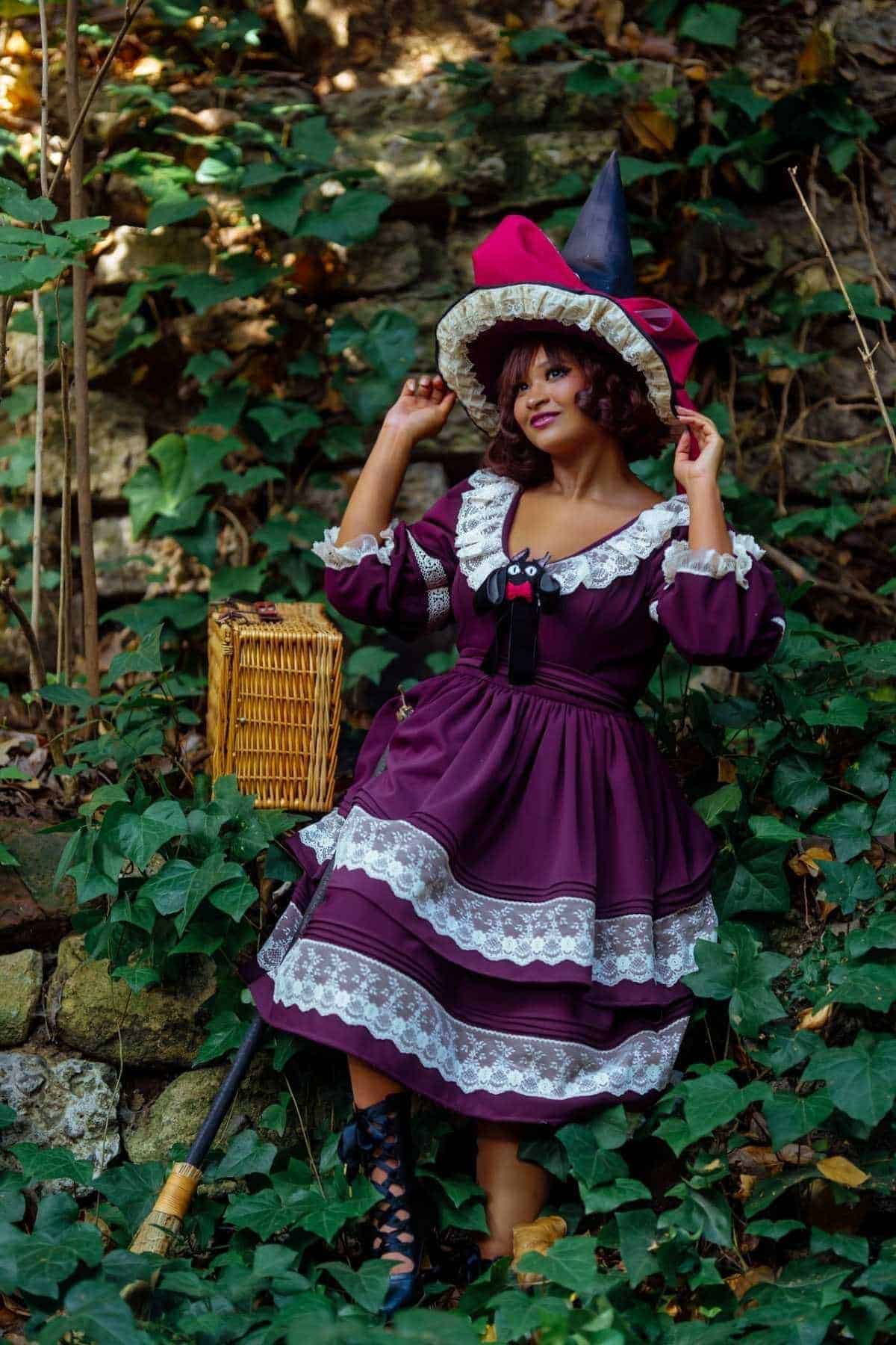 A person in a purple dress and witch hat stands outdoors among ivy, holding their hat, with a wicker basket and broom nearby.