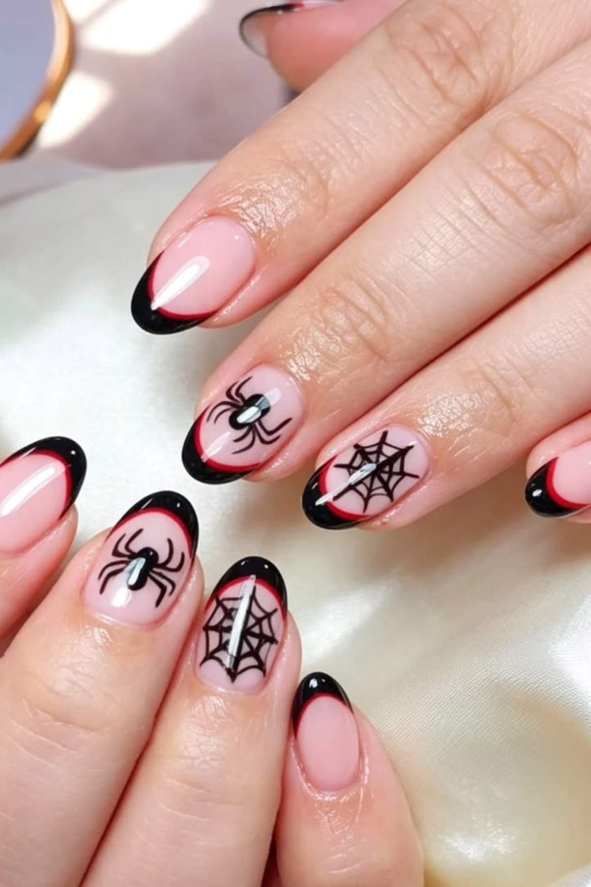 Hands with almond-shaped nails featuring black French tips, red accents, and Halloween-themed spider and spiderweb nail art on a light pink base.