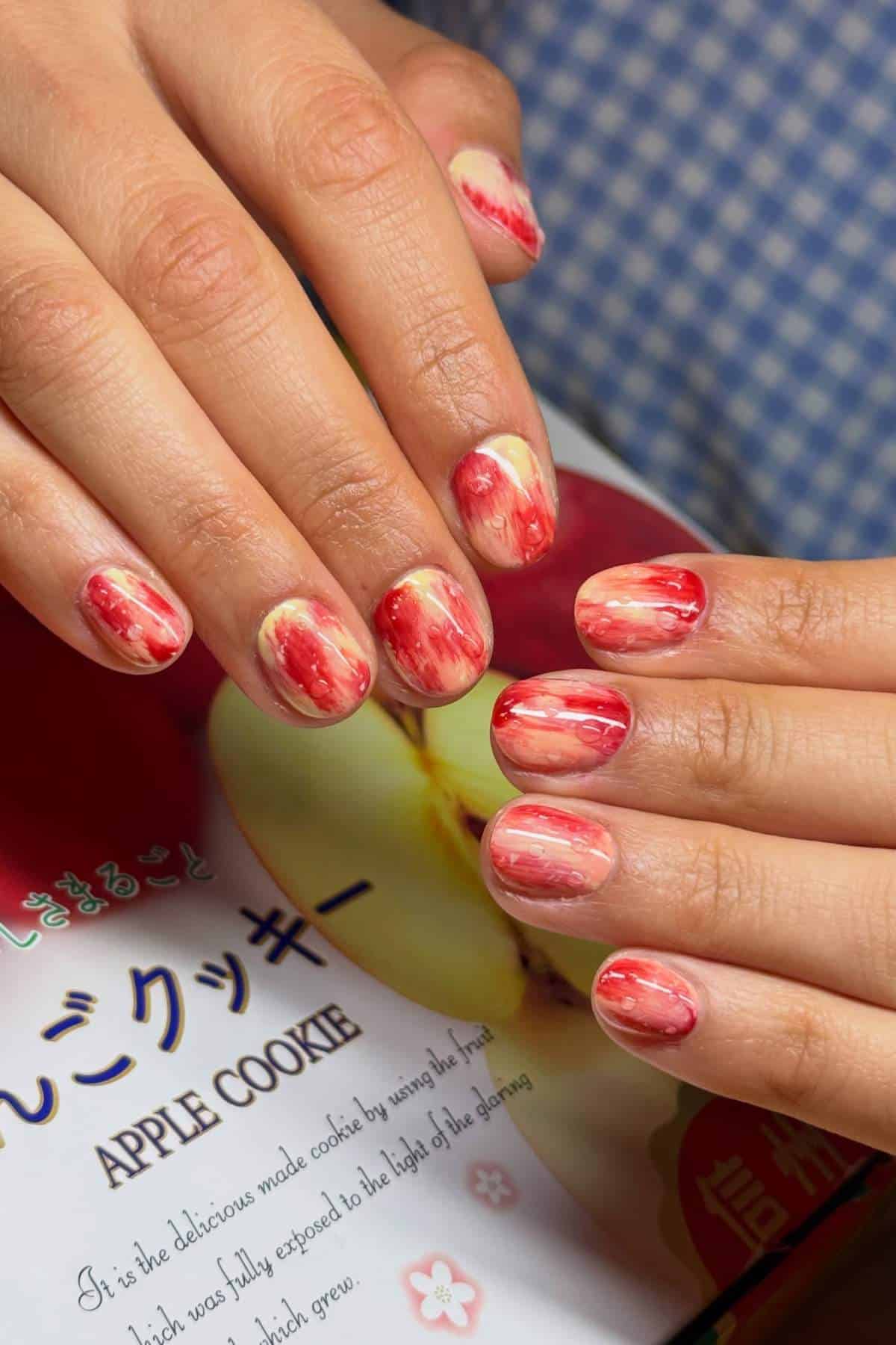 Hands with short, rounded nails painted in a red and cream marbled pattern, resting on a book or packaging labeled "Apple Cookie" with Japanese and English text.