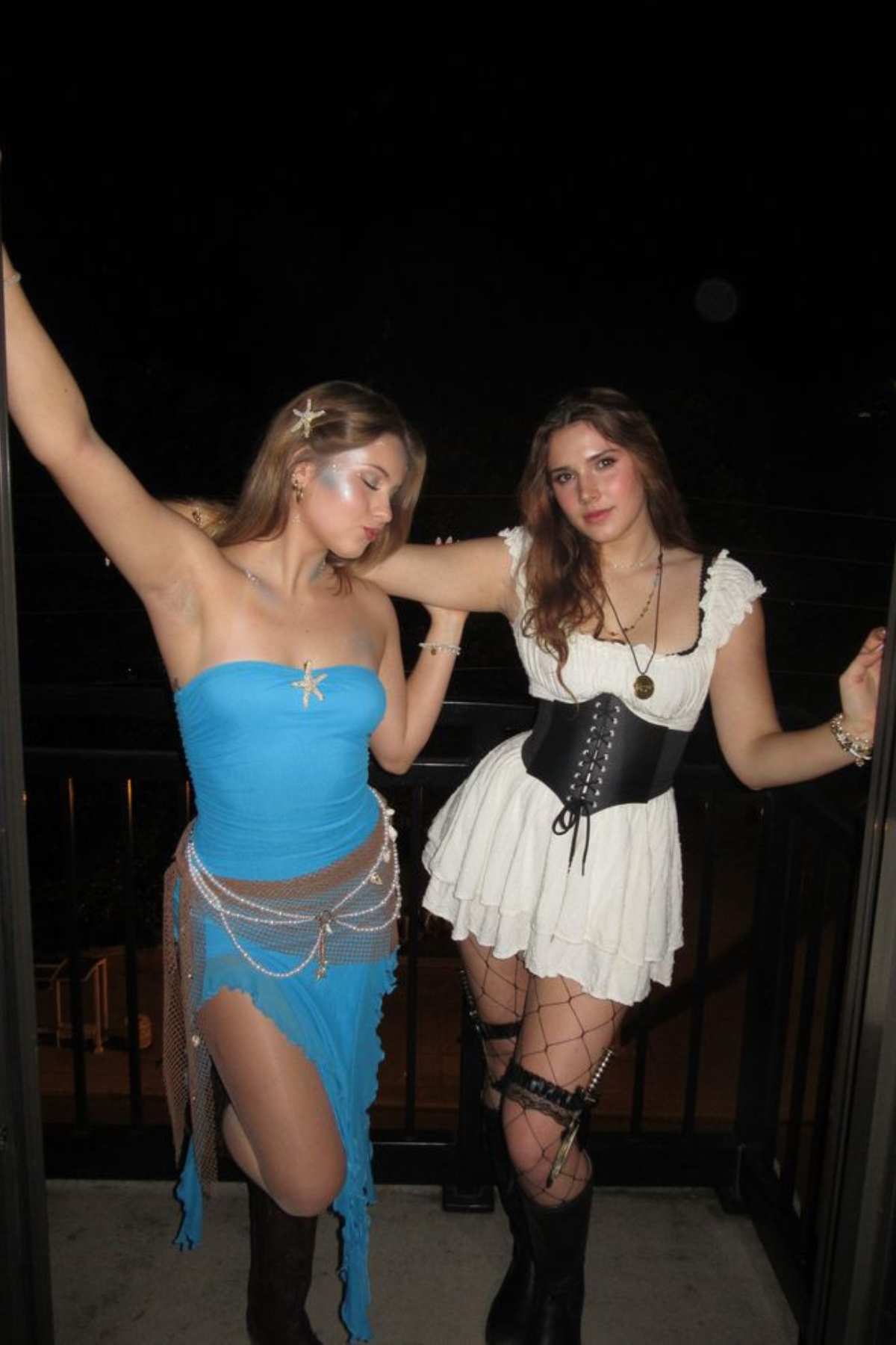 Two women in costumes pose on a balcony at night; one wears a blue dress with accessories, and the other wears a white dress with a black corset, boots, and netted leg wraps.