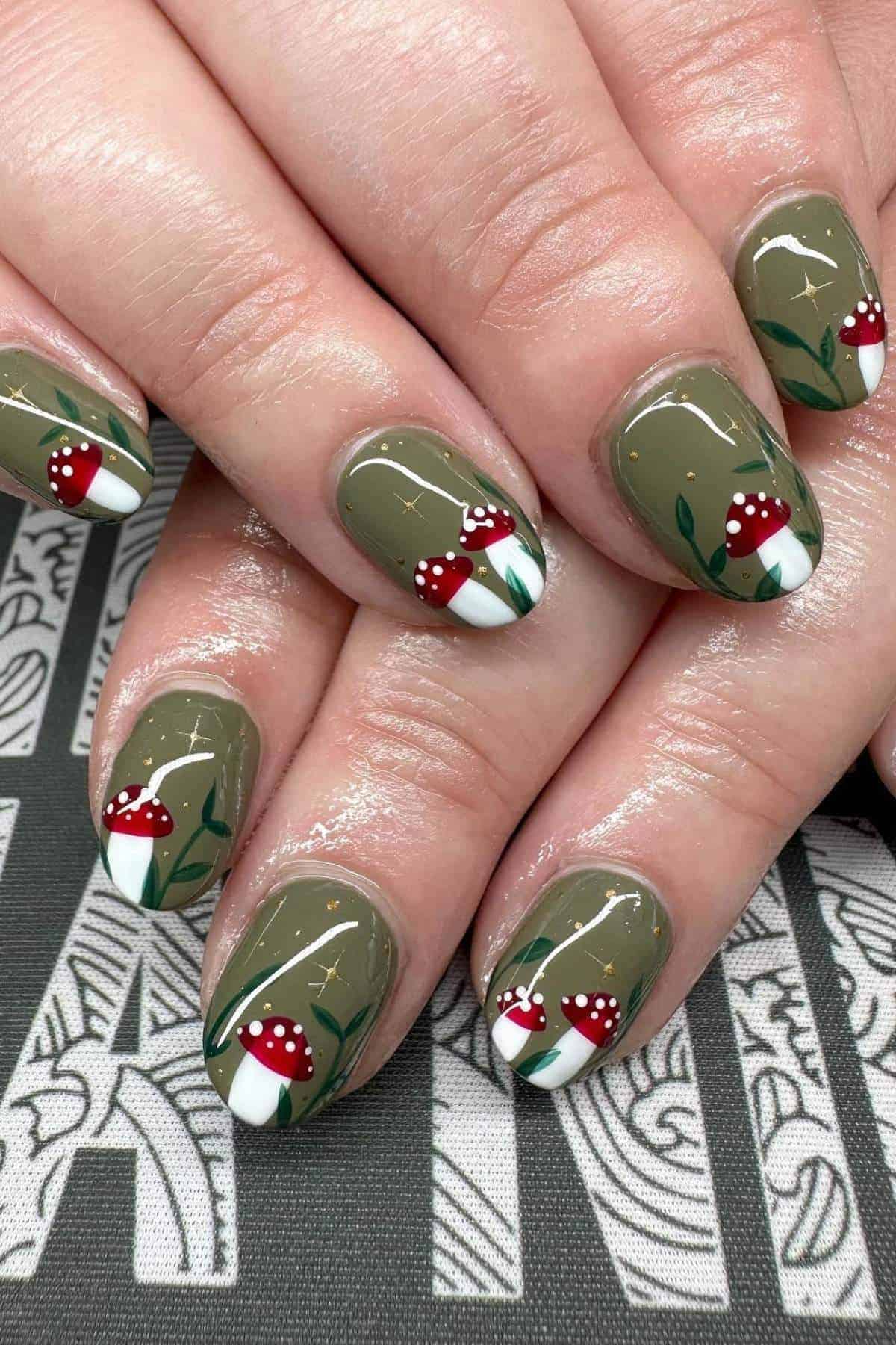 Close-up of manicured hands with olive green nails, featuring painted red and white mushrooms and small green leaf designs on each nail.