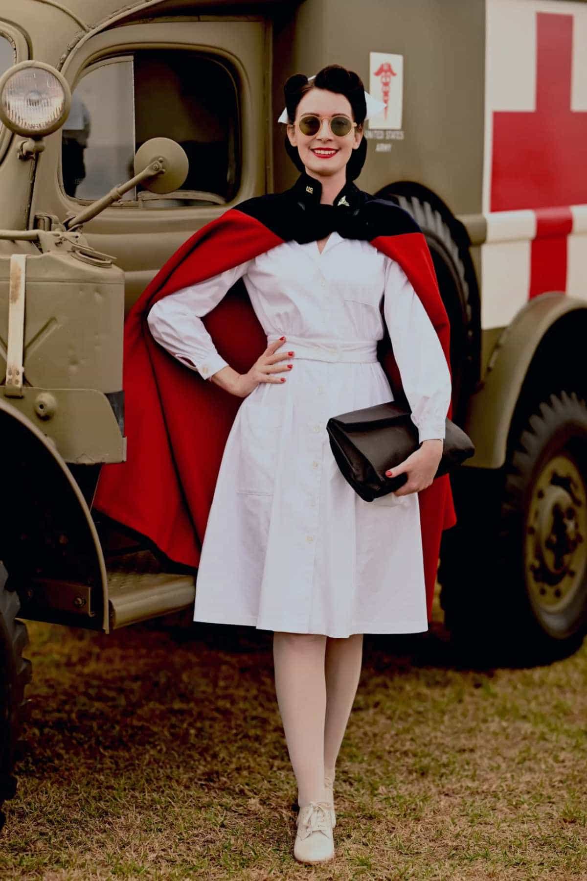 A woman in a vintage nurse uniform and red cape stands in front of a military ambulance with a red cross symbol, holding a black clutch bag.